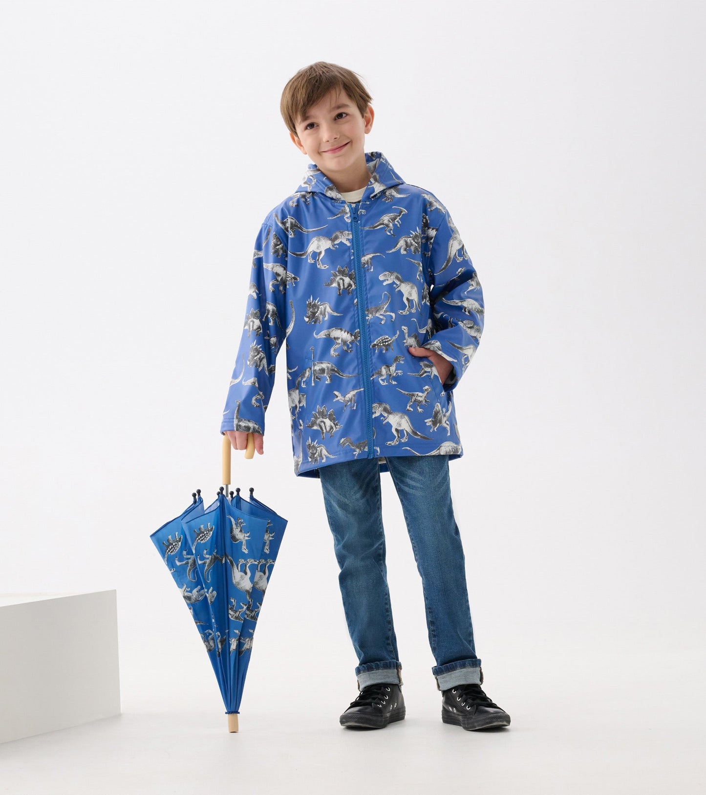 Blue zip-up rain jacket featuring dinosaur patterns, paired with a matching umbrella, worn by a smiling boy.