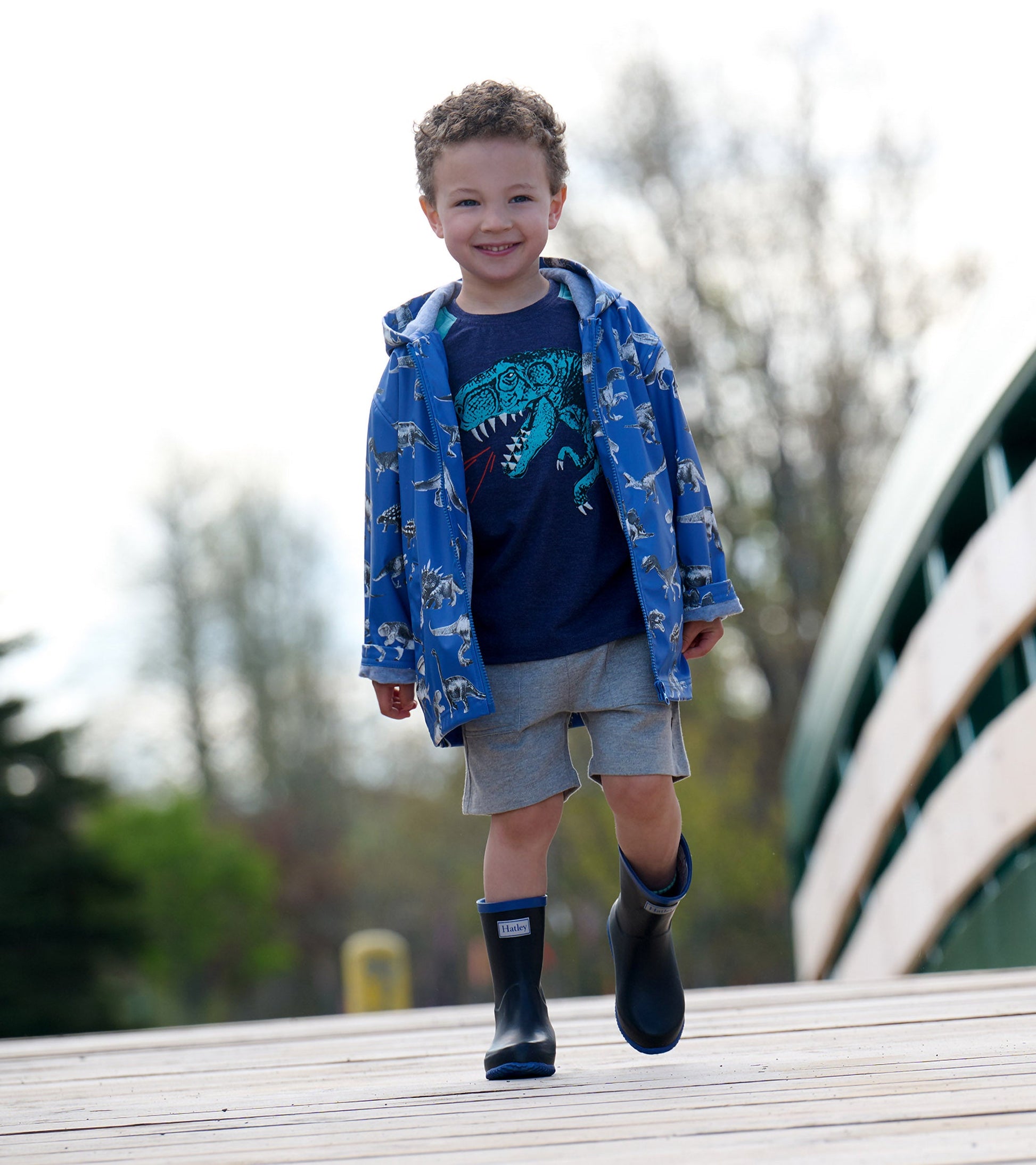 Blue zip-up rain jacket with dinosaur print, worn by a smiling boy, paired with gray shorts and black rain boots.