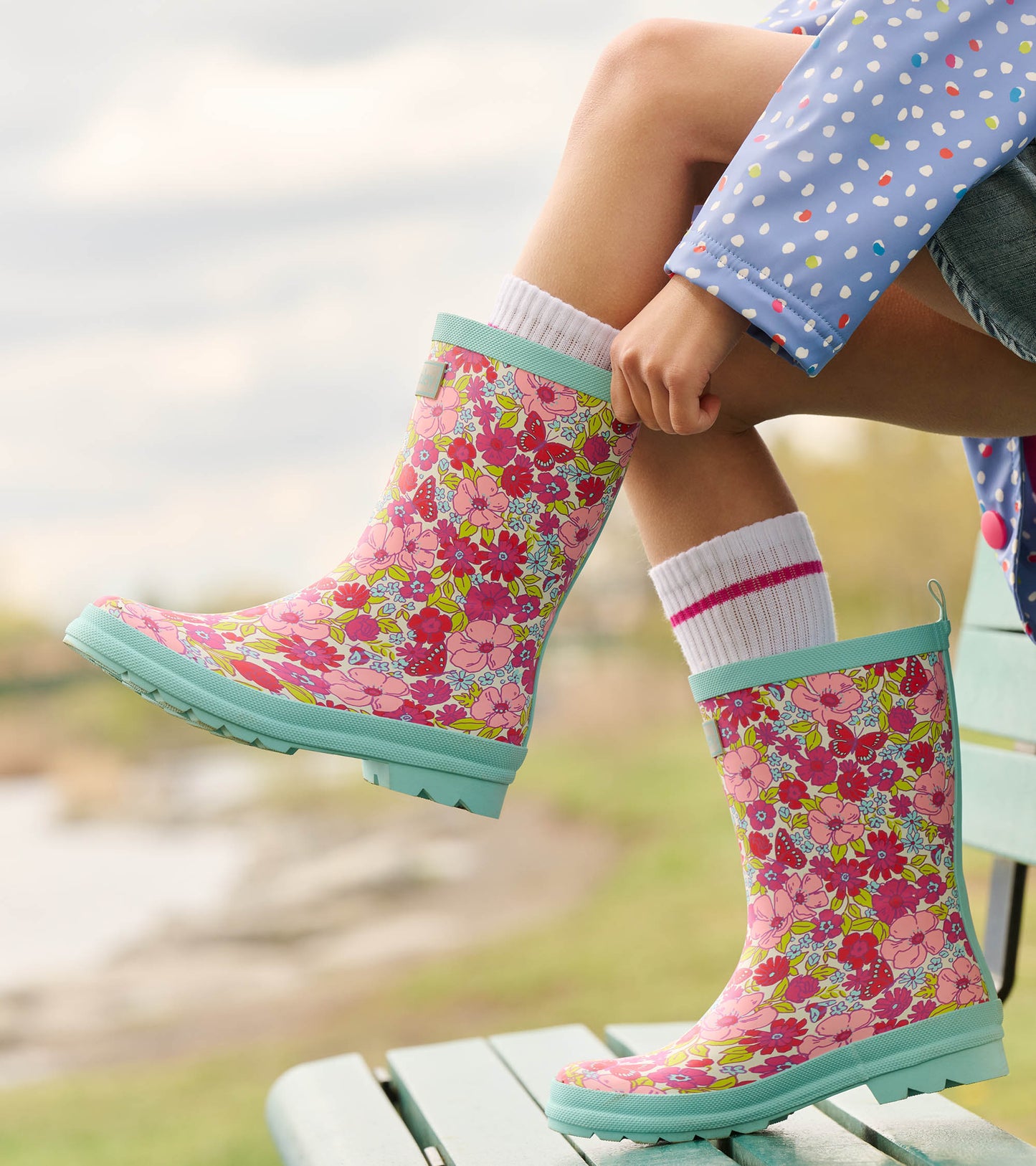 Pink matte rain boots with colorful floral pattern and mint green soles, worn with white socks, on a wooden bench.