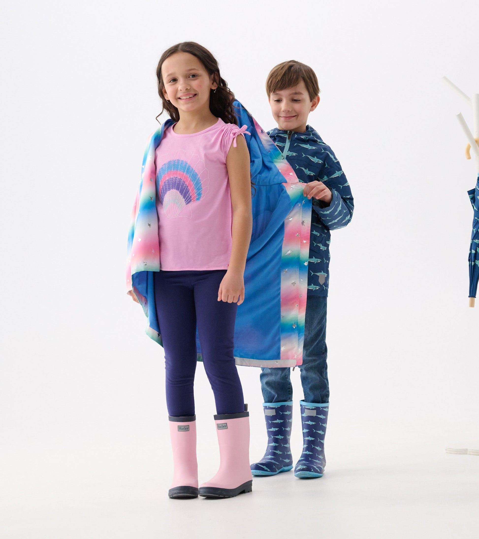 A girl in a pink top and leggings wears a colorful glitter star A-line swing jacket, paired with pink rain boots.