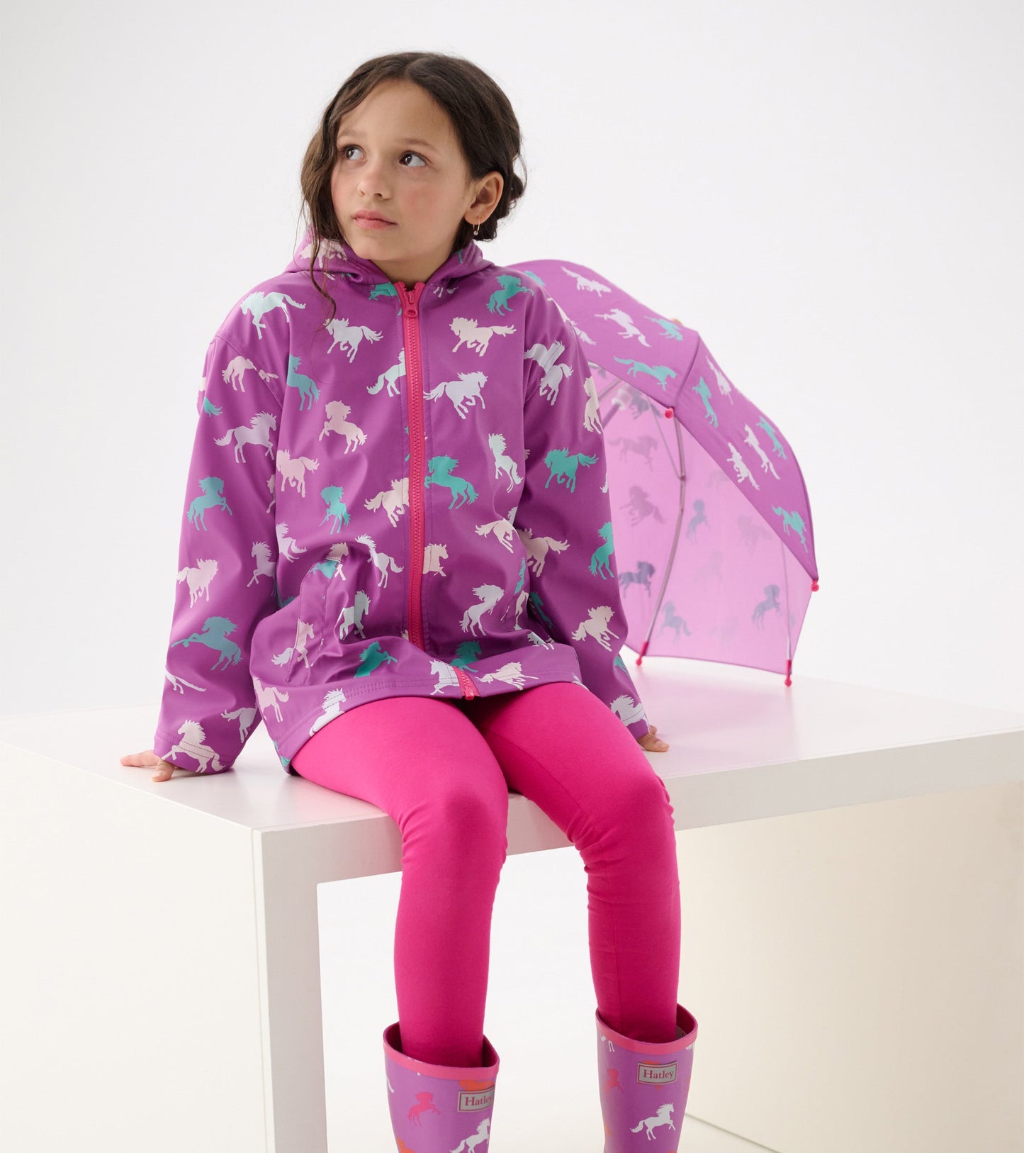 Purple zip-up rain jacket with horse pattern, paired with pink leggings and boots, worn by a girl sitting on a white surface.