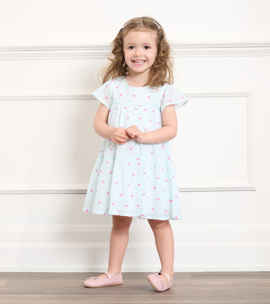 Light blue woven dress with pink heart pattern, short flutter sleeves, and a tiered skirt, worn by a smiling girl.