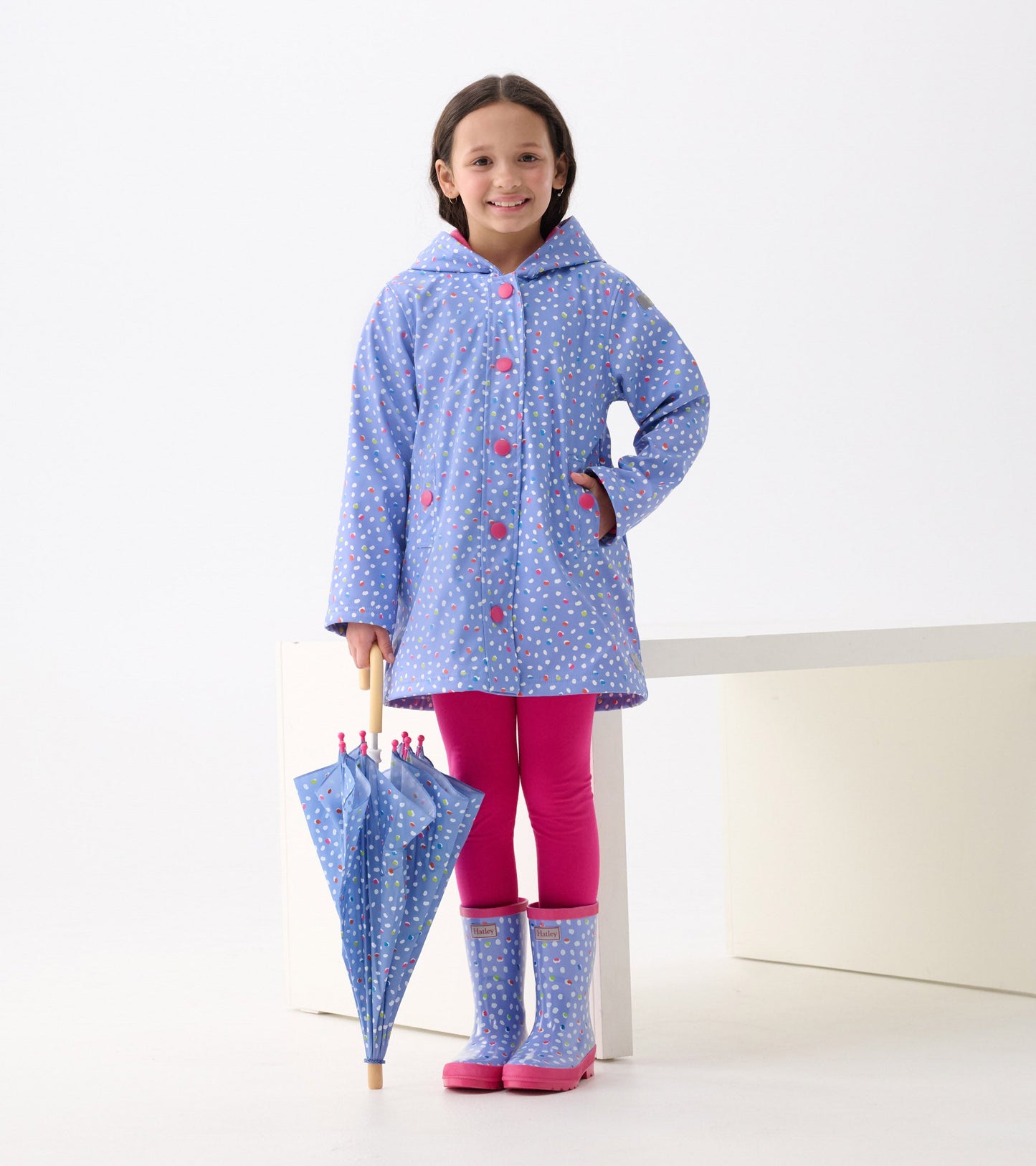 Shiny blue rain boots with colorful polka dots and pink soles, worn by a girl in a matching raincoat and pink leggings.