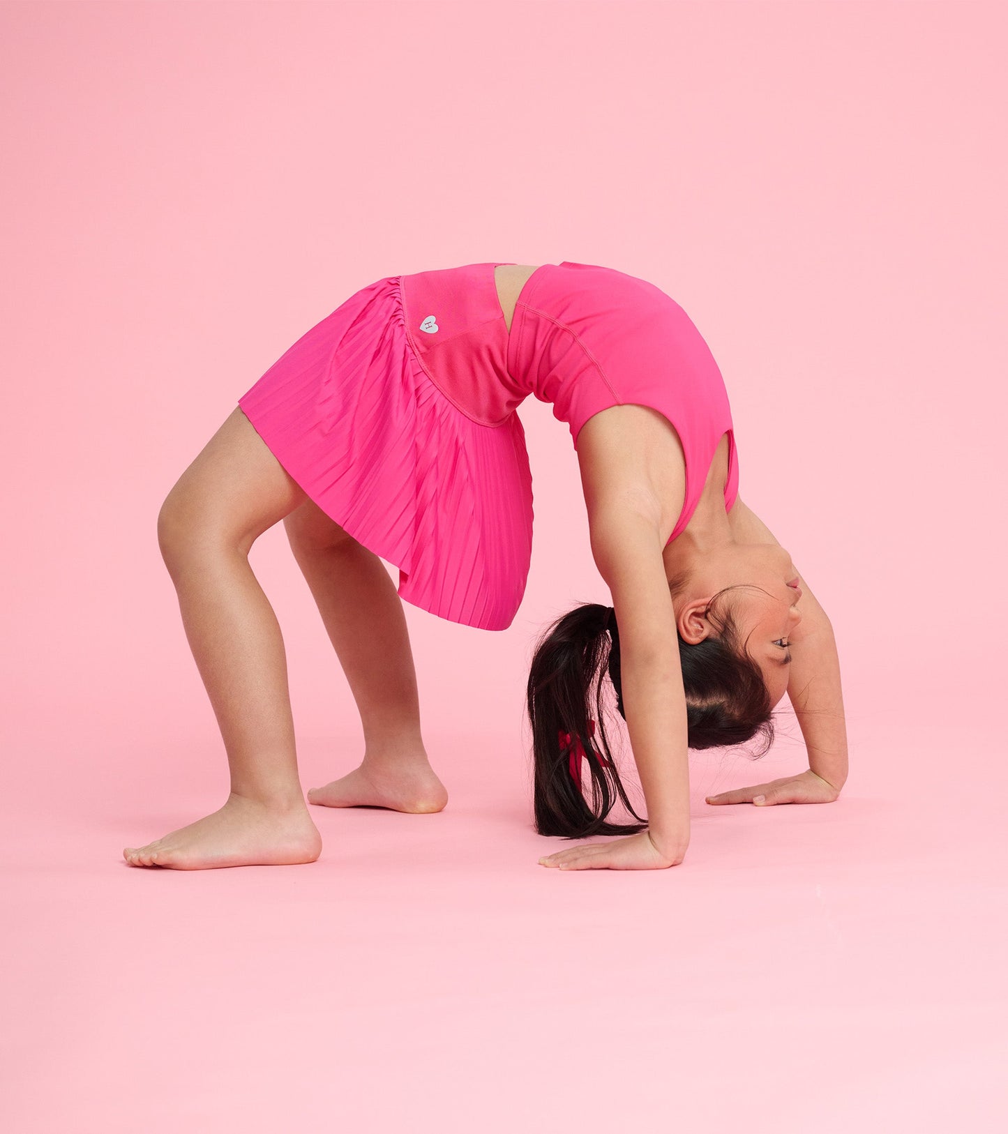 Bright pink sporty skort with pleated design, worn by a girl performing a backbend on a pink background.