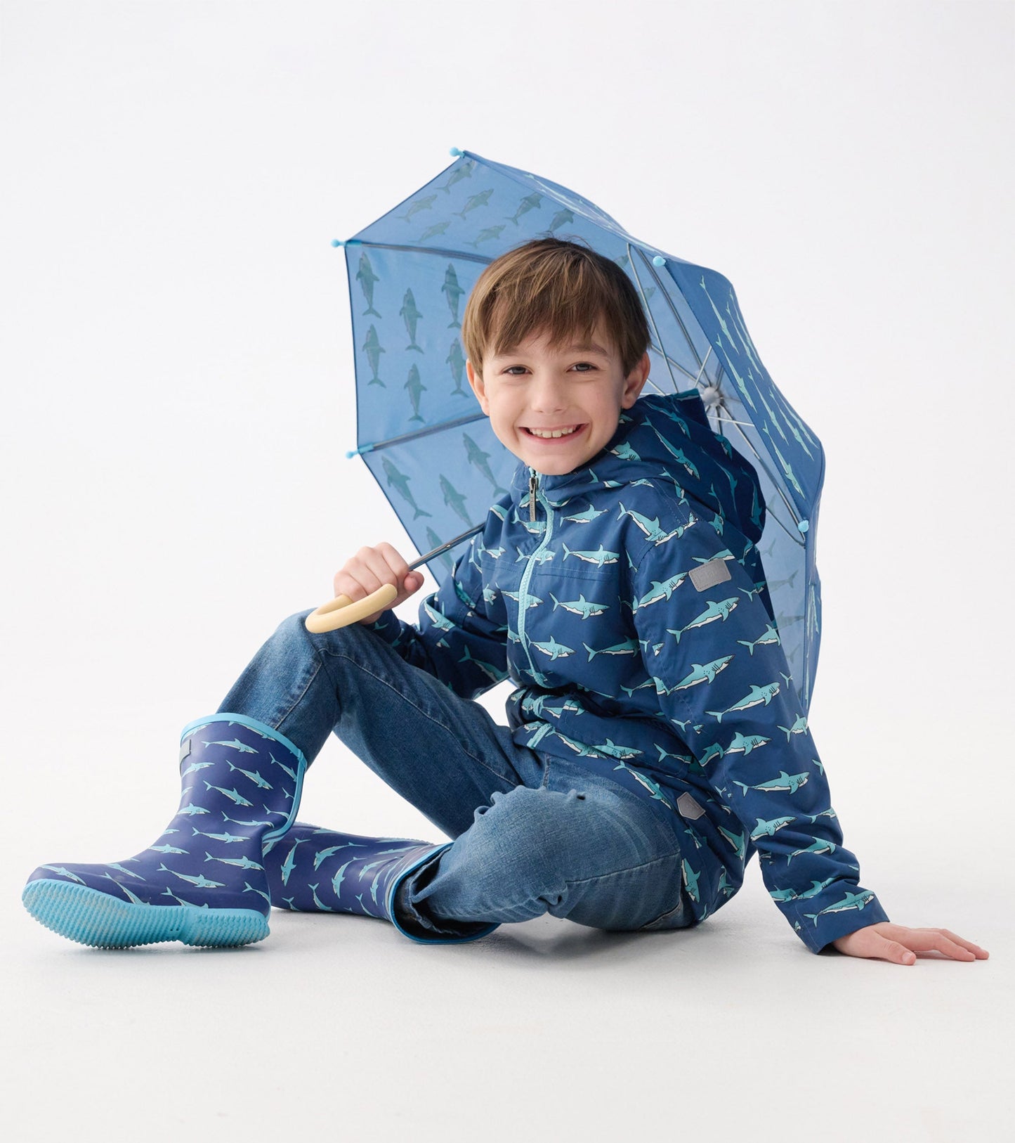 Lightweight blue rain jacket with shark pattern, paired with matching rain boots, worn by a smiling boy holding an umbrella.