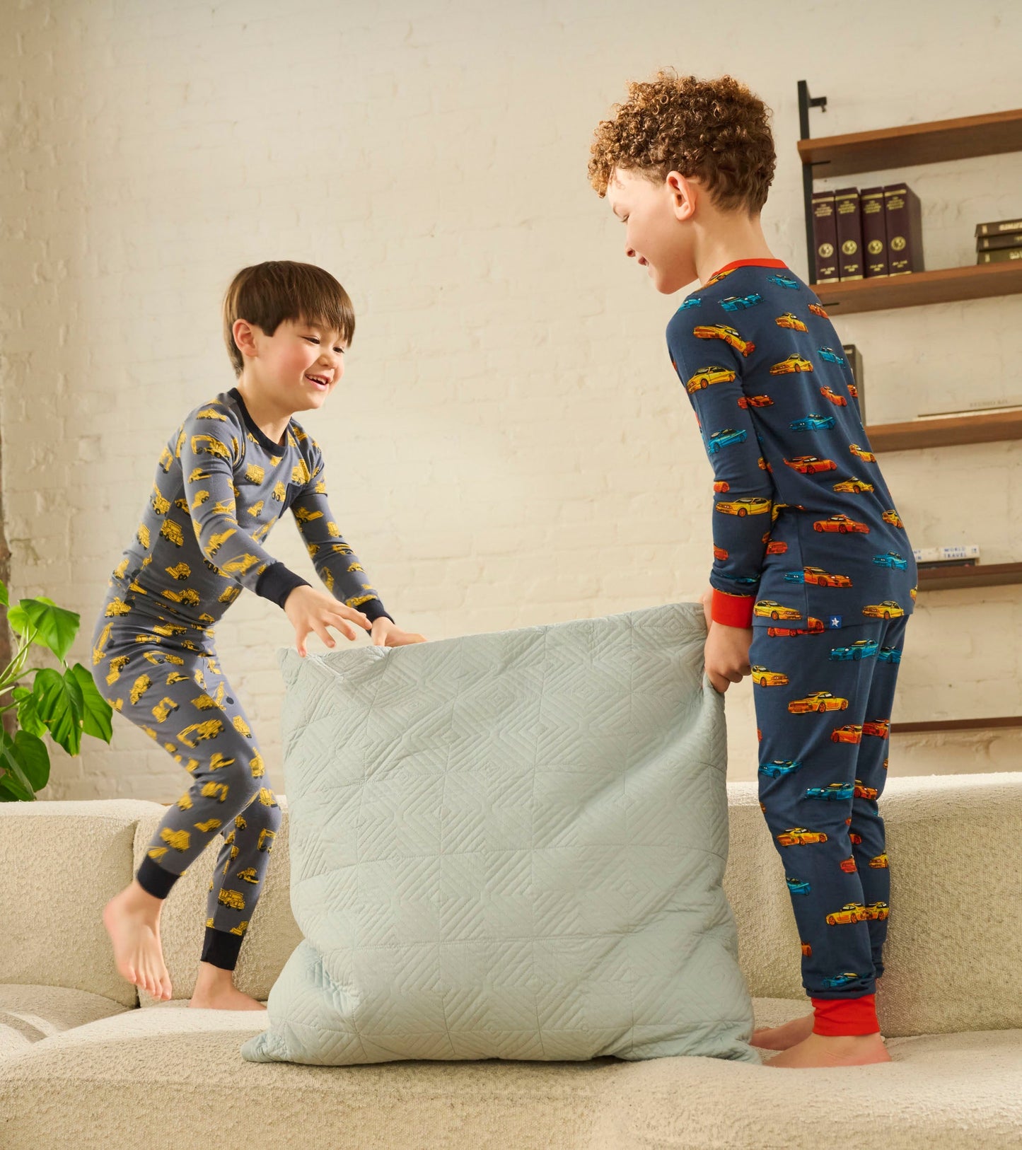 Two boys play on a couch, one in gray pajamas with yellow trucks, the other in navy pajamas with colorful cars.