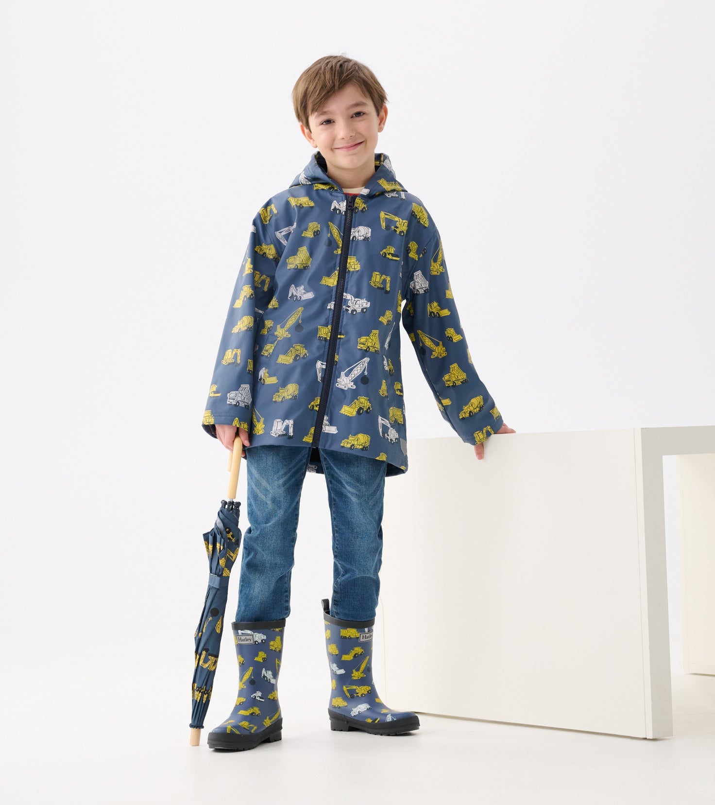 Blue zip-up rain jacket with construction vehicle patterns, paired with jeans and matching rain boots, held umbrella.
