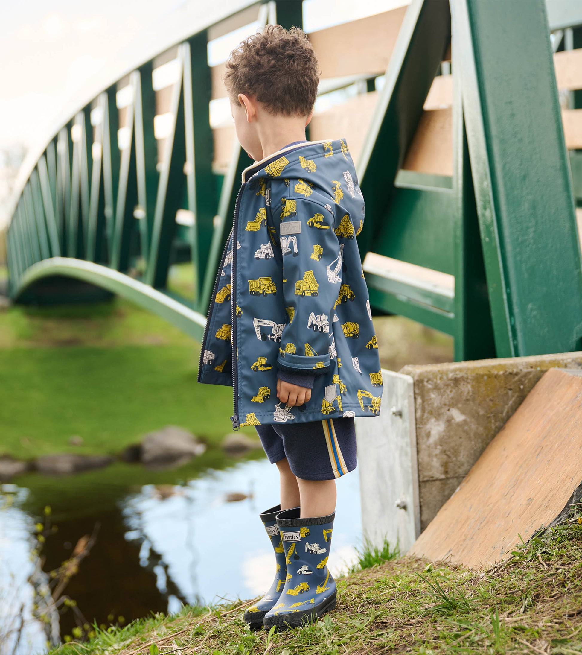 Boys' zip-up rain jacket in blue with yellow construction vehicle print, worn with shorts and rain boots, standing by a pond.