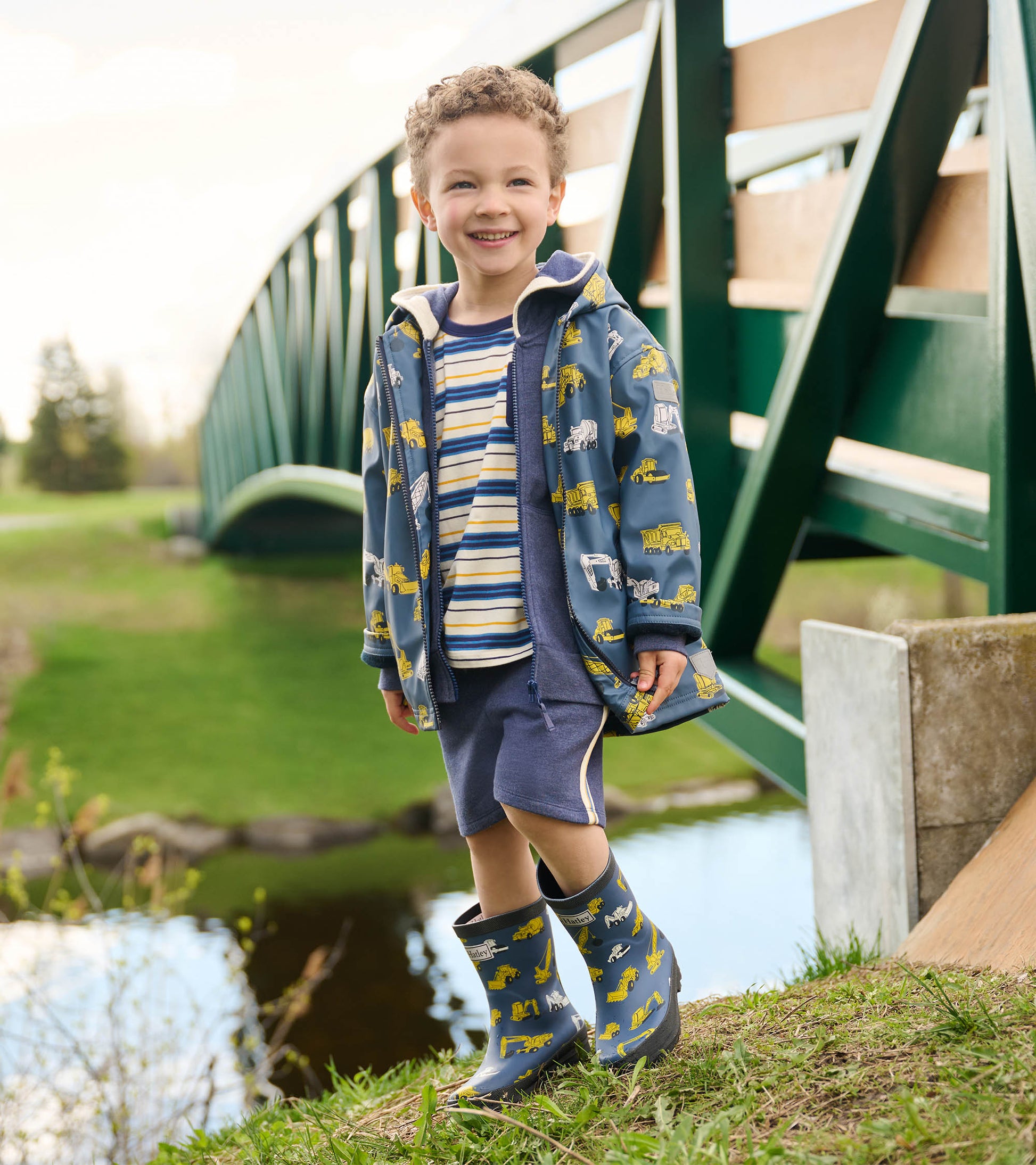 Blue zip-up rain jacket with construction vehicle prints, worn over a striped shirt and shorts, paired with matching rain bo…