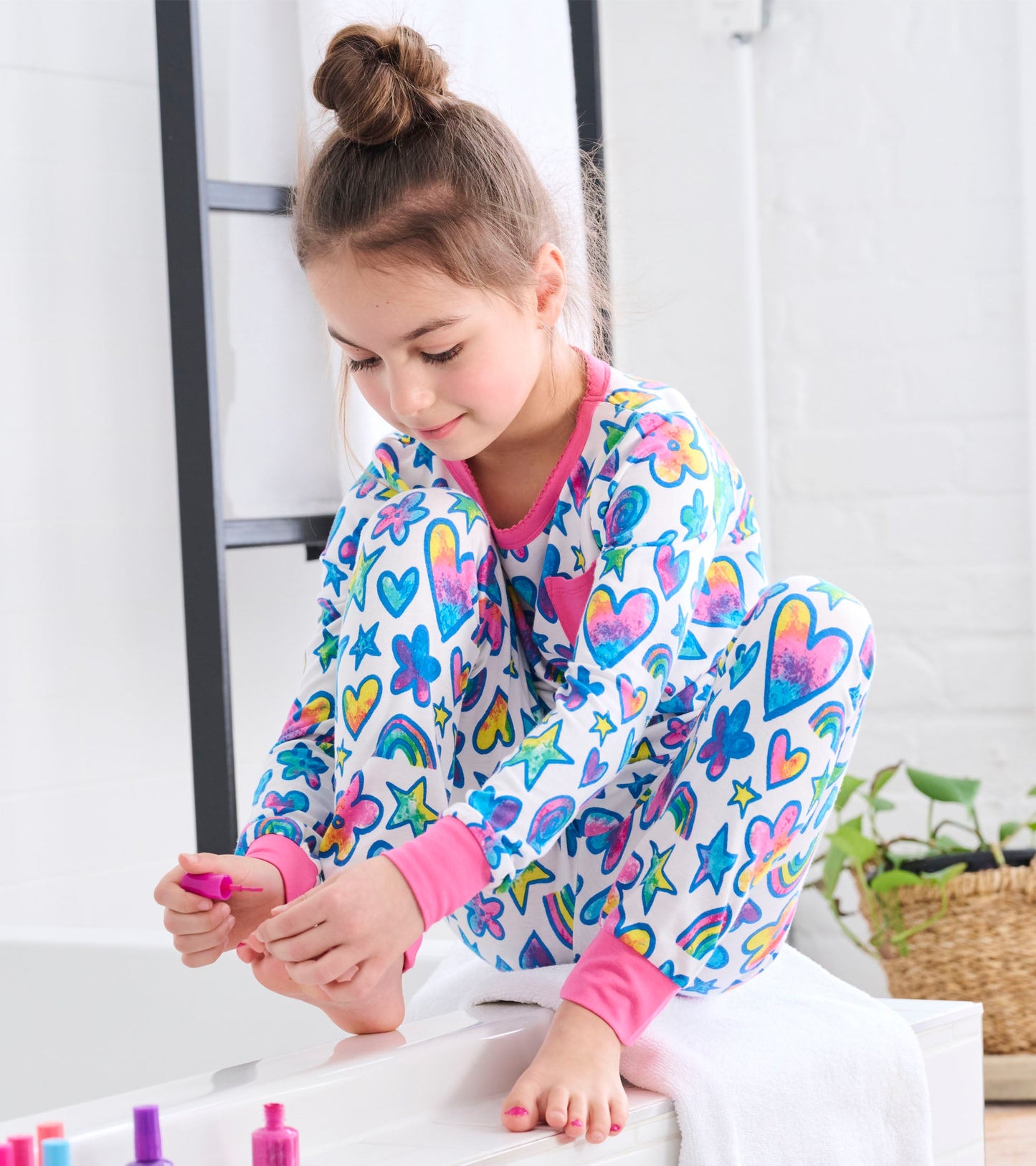 Colorful pajama set featuring watercolour hearts, stars, and flowers, with pink cuffs, worn by a girl sitting on a tub edge.