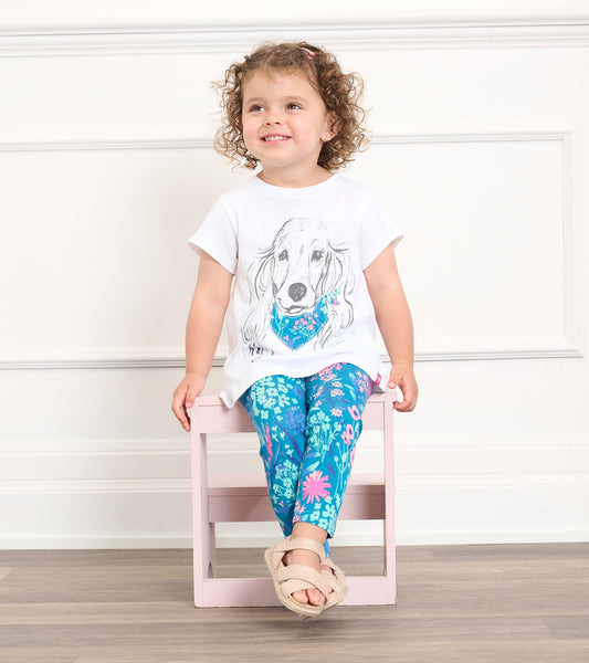 White graphic tee featuring a puppy illustration, paired with blue floral leggings, worn by a smiling toddler.
