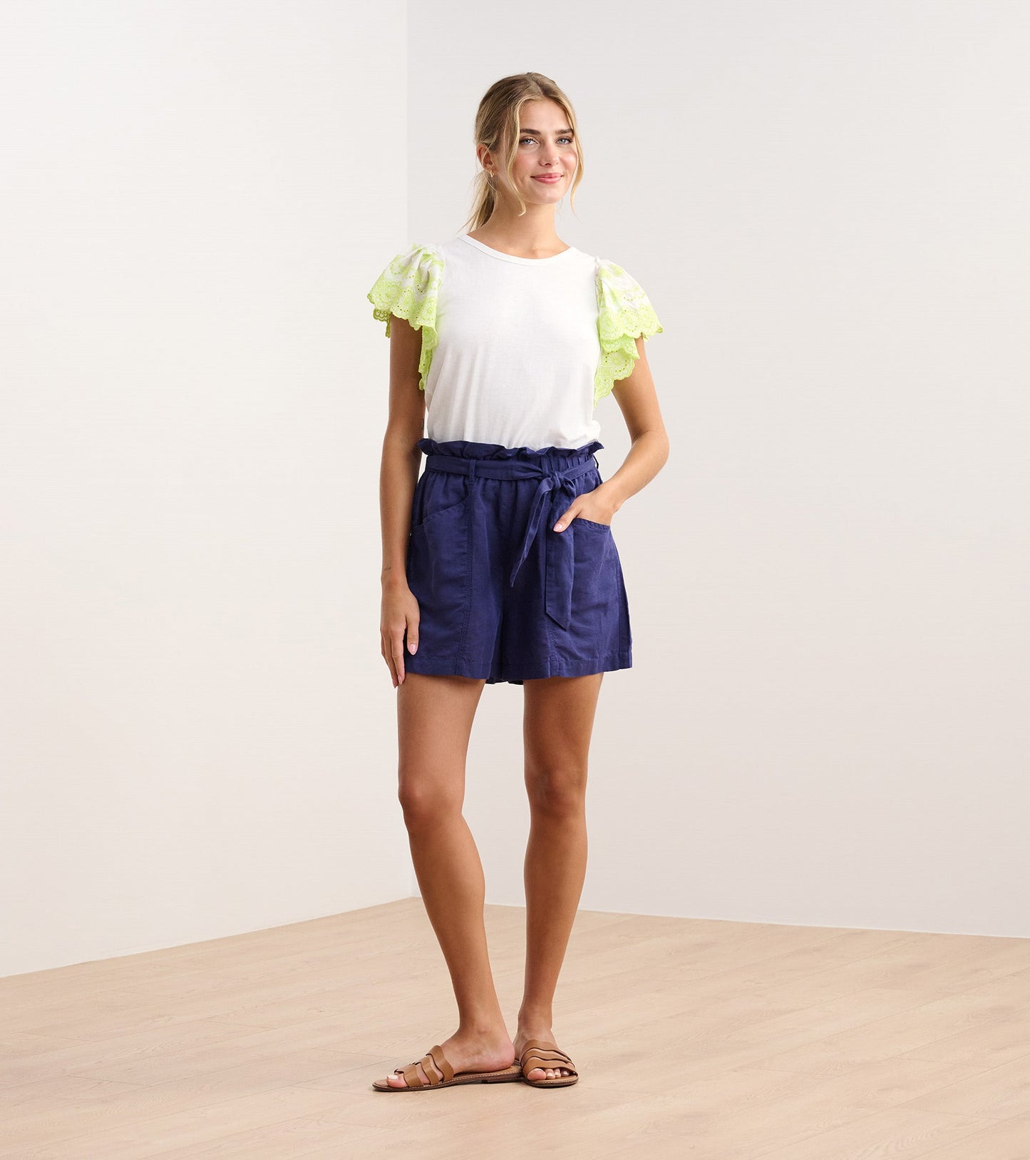 White tank top with eyelet sleeves, paired with navy shorts, worn by a model standing on a wooden floor.