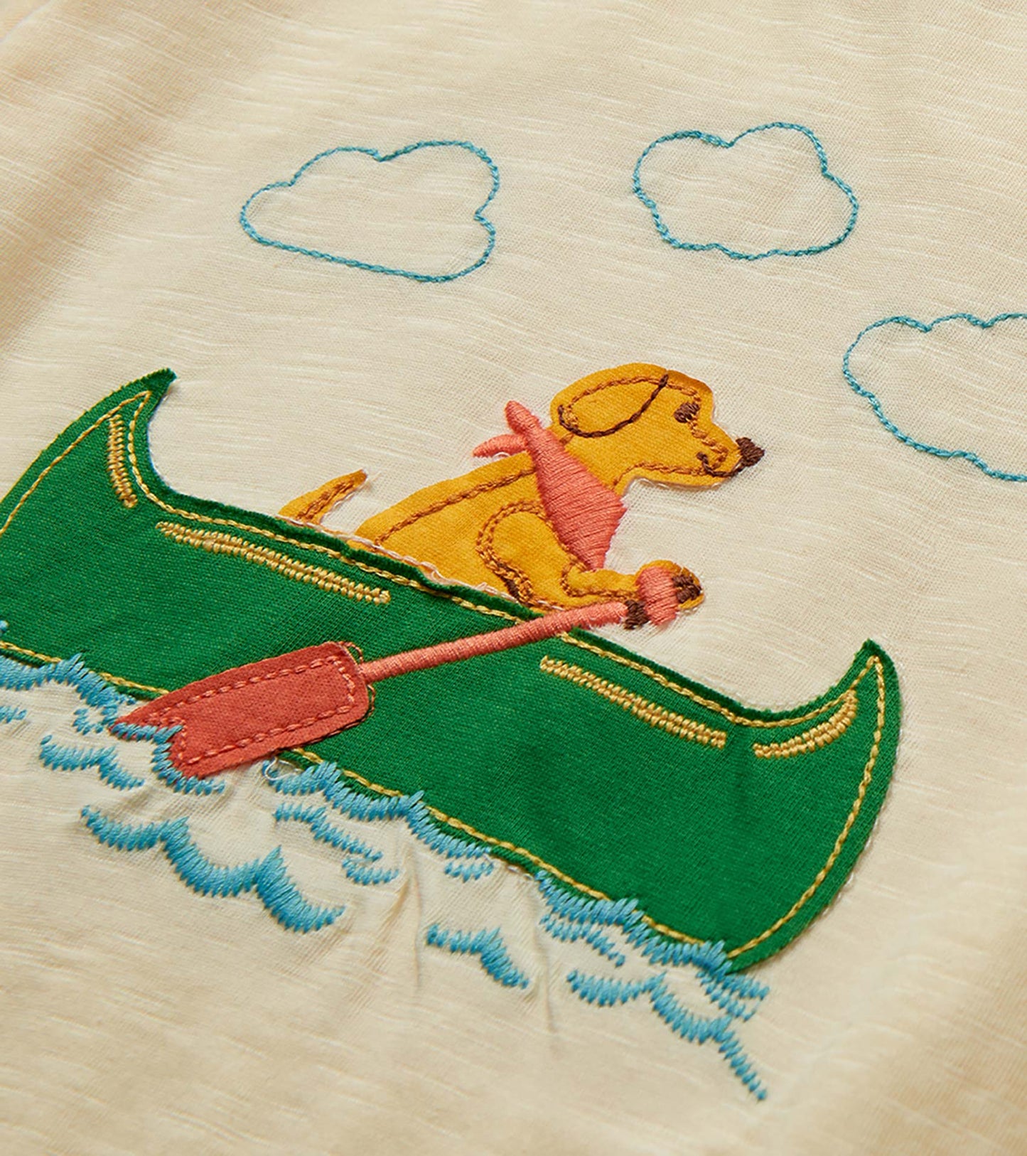 Close-up of cream T-shirt with embroidered dog in green canoe on blue waves and cloud outlines above
