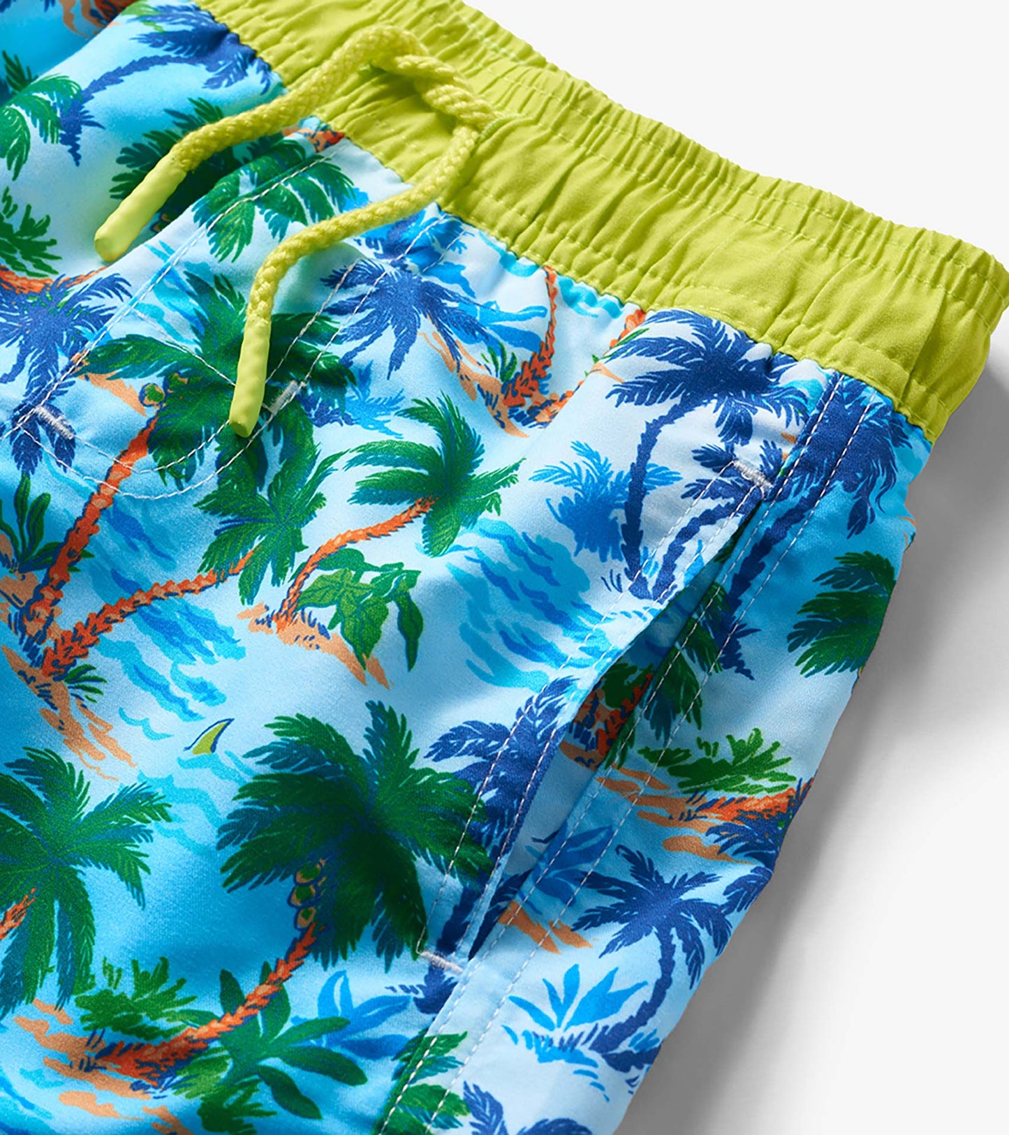 Blue boys swim trunks with tropical palm print, lime green waistband, and drawstring detail.