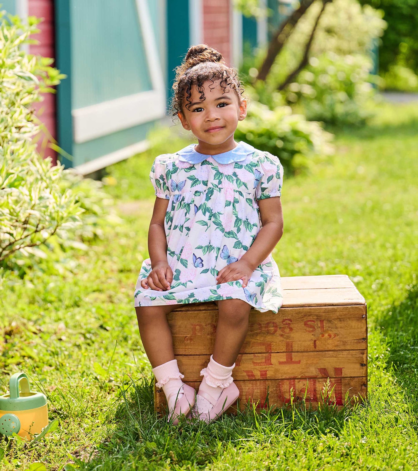 Toddler girl seated outdoors in the floral butterfly dress with blue Peter Pan collar, front view