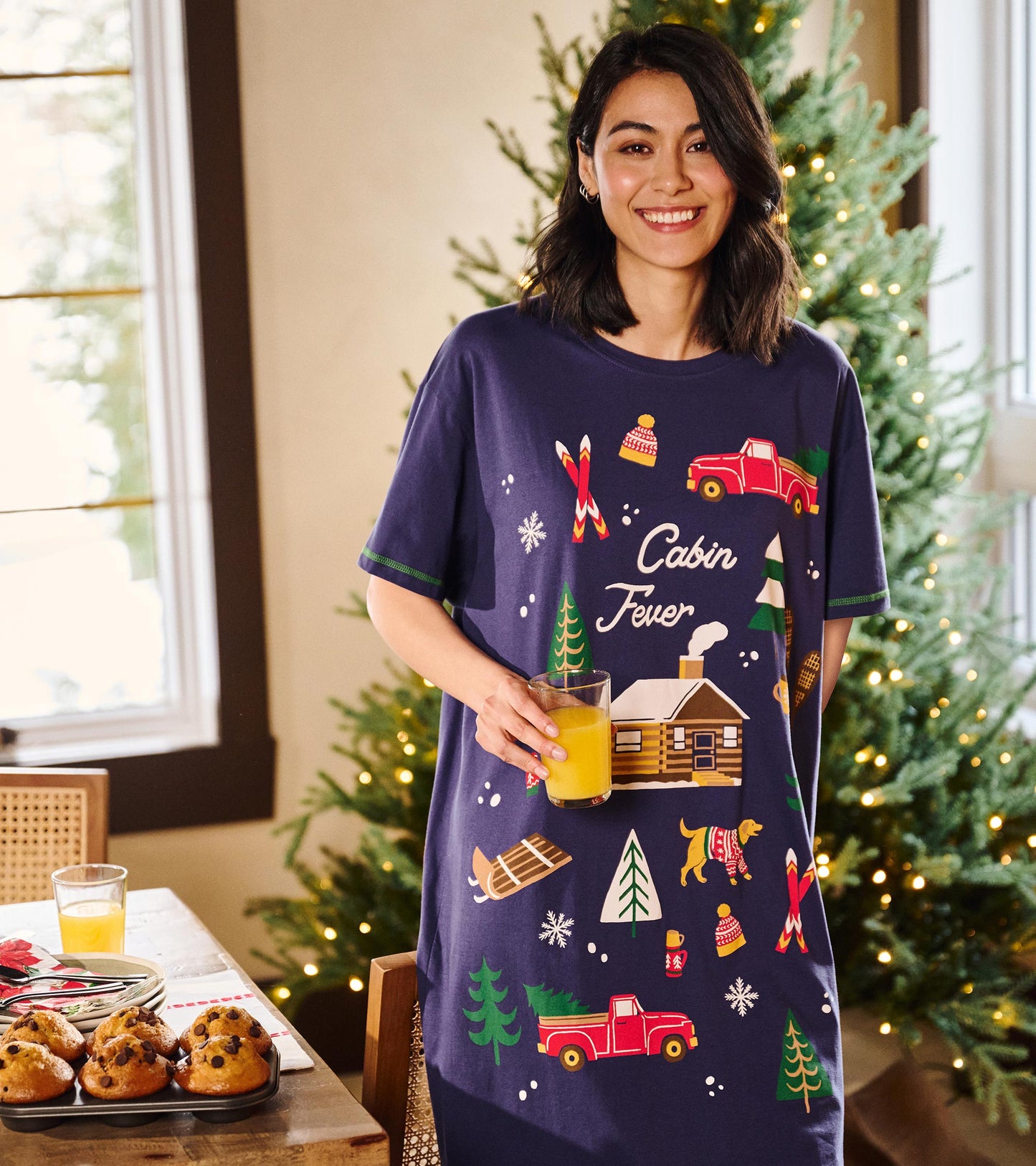 Model wearing a navy cabin-print sleepshirt with short sleeves beside a lit Christmas tree