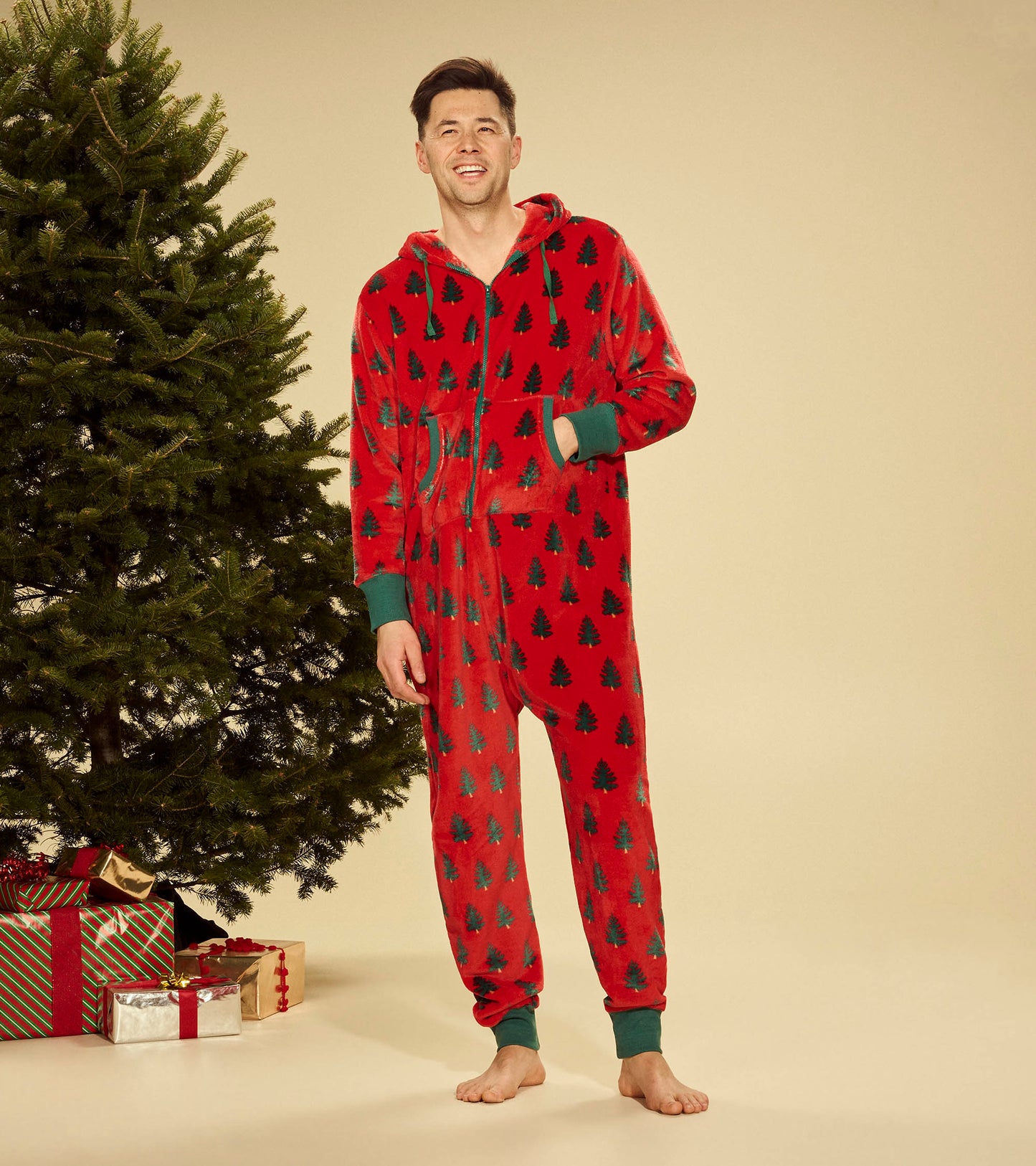 Red fleece jumpsuit featuring a tree pattern, with a hood and green cuffs, worn by a smiling man next to a Christmas tree.