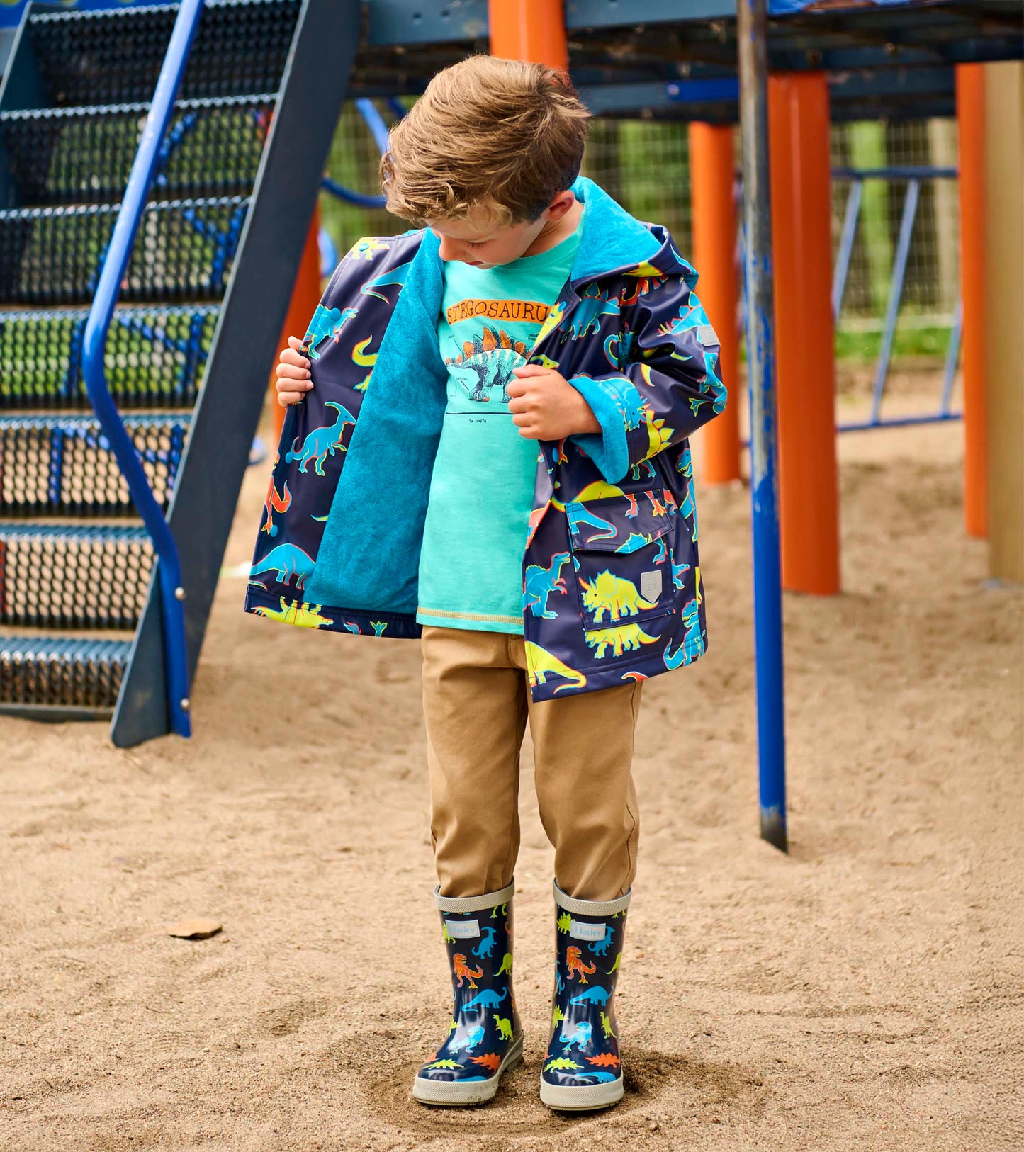Boy wearing a navy dinosaur-print rain jacket with teal lining, open front, and hood at a playground