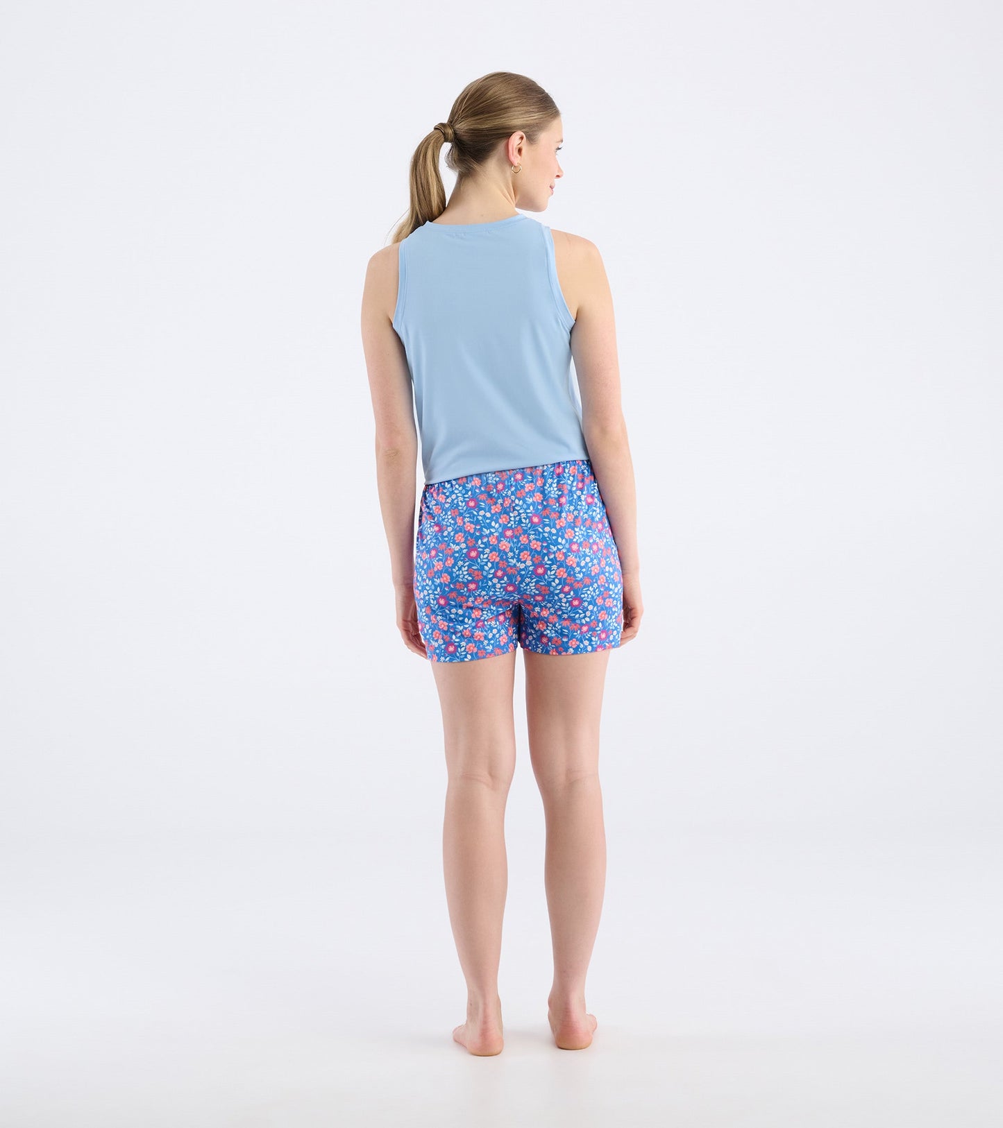 Back view of a woman wearing blue pajama shorts with a colorful floral pattern and a light blue tank top.