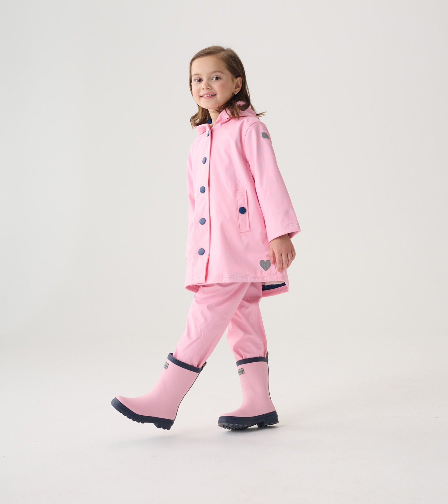 Pink button-up rain jacket with navy buttons, paired with matching pink rain pants and boots, worn by a smiling girl.