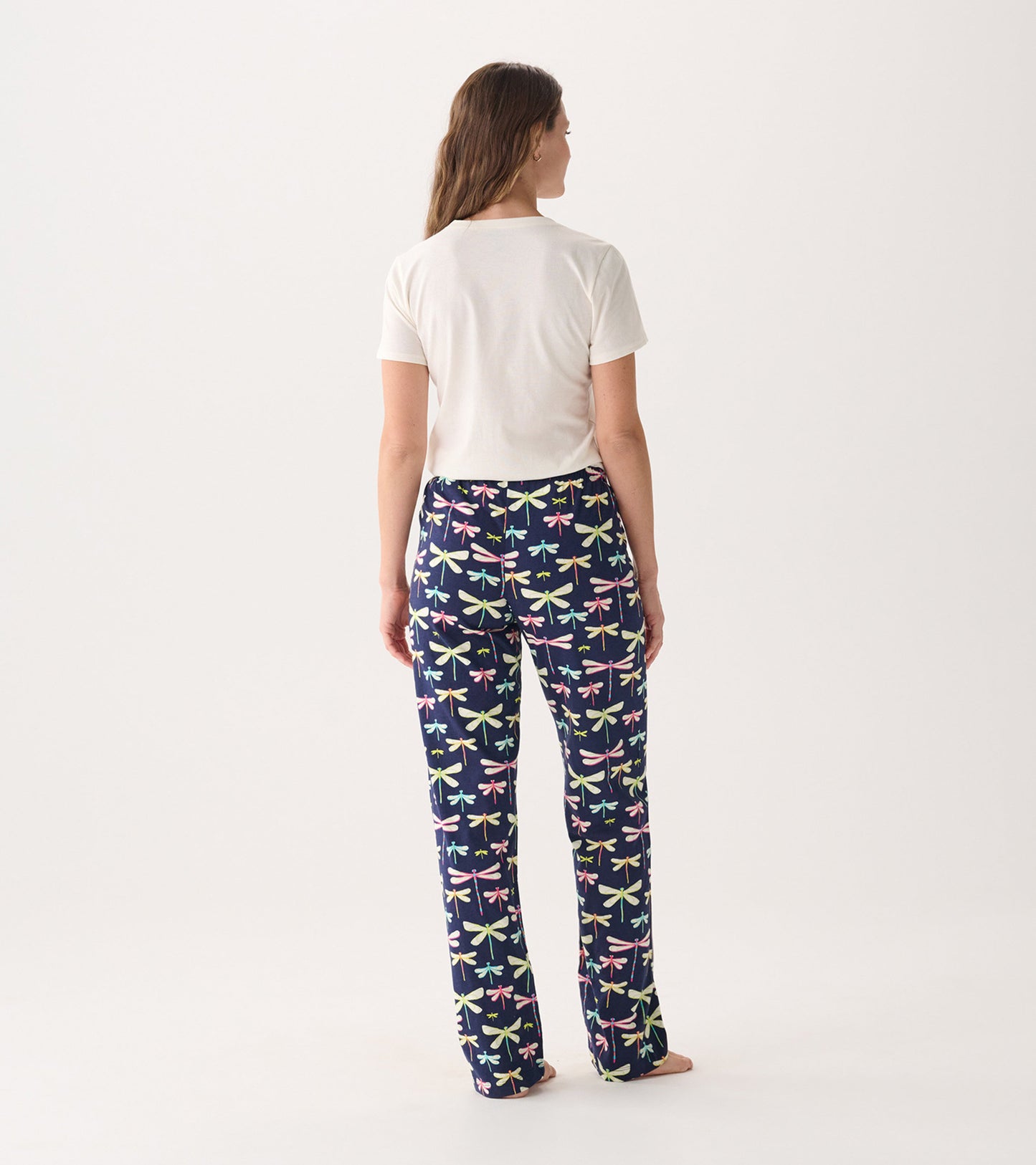 Back view of a woman wearing a cream short-sleeve pajama t-shirt with colorful dragonfly-patterned pants.