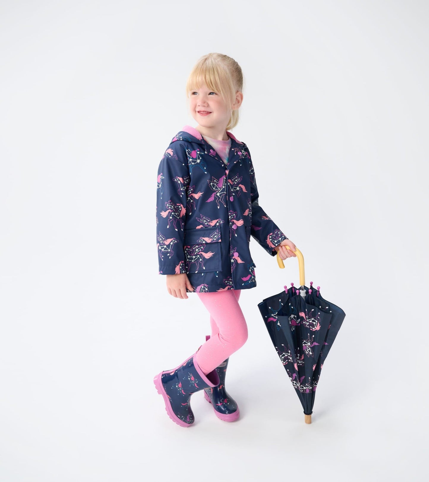 Navy rain jacket with unicorn constellations, pink accents, paired with pink leggings and matching rain boots, holding an um…