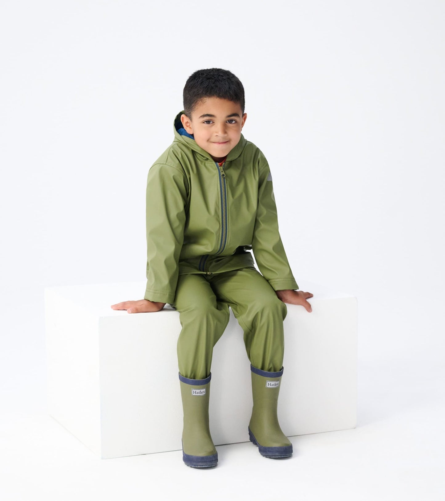 Forest green matte rain boots with a blue trim, designed for kids, shown on a child sitting on a white block.
