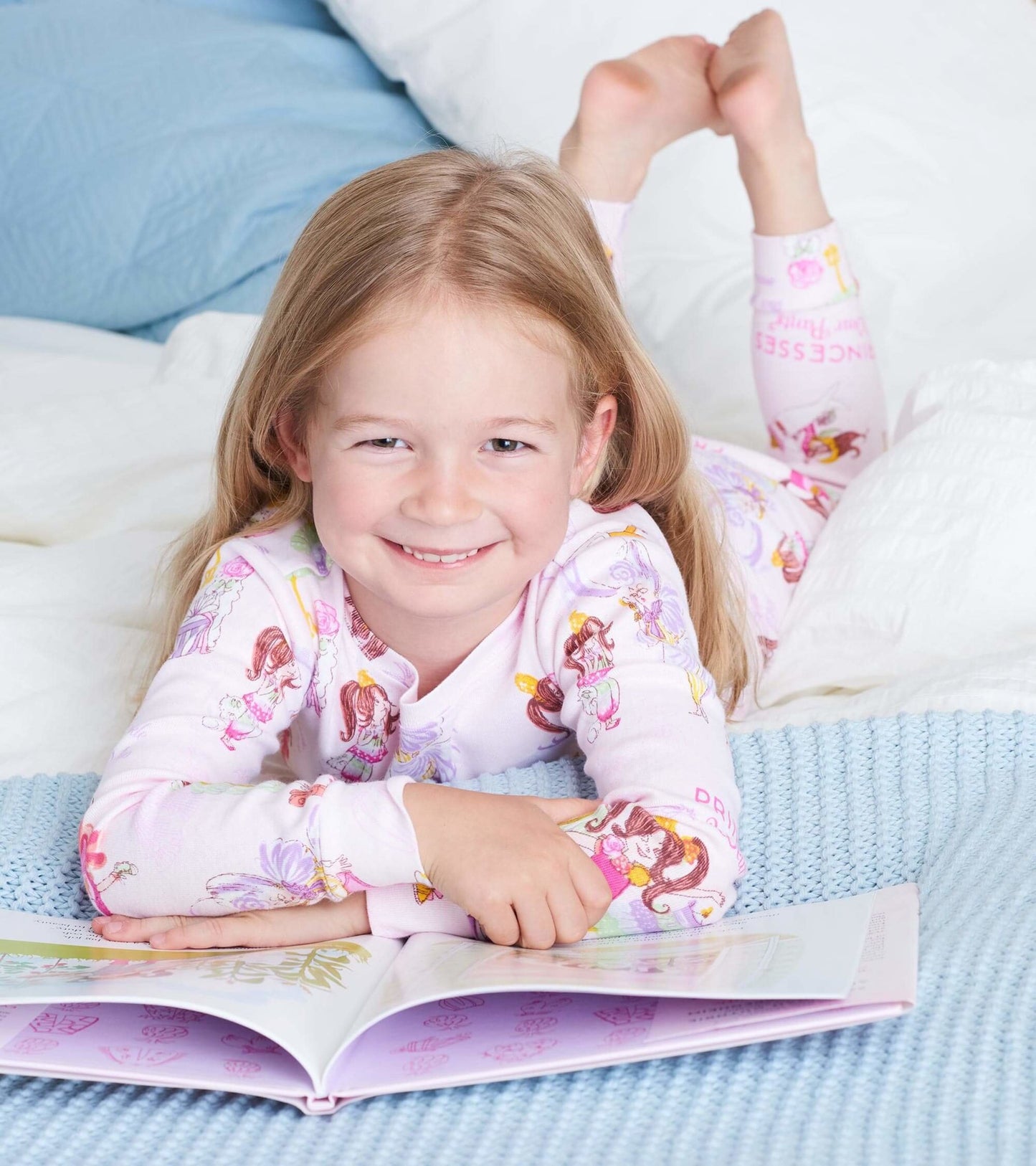 Light pink pajama set featuring princess illustrations, worn by a smiling girl reading on a bed with blue and white bedding.