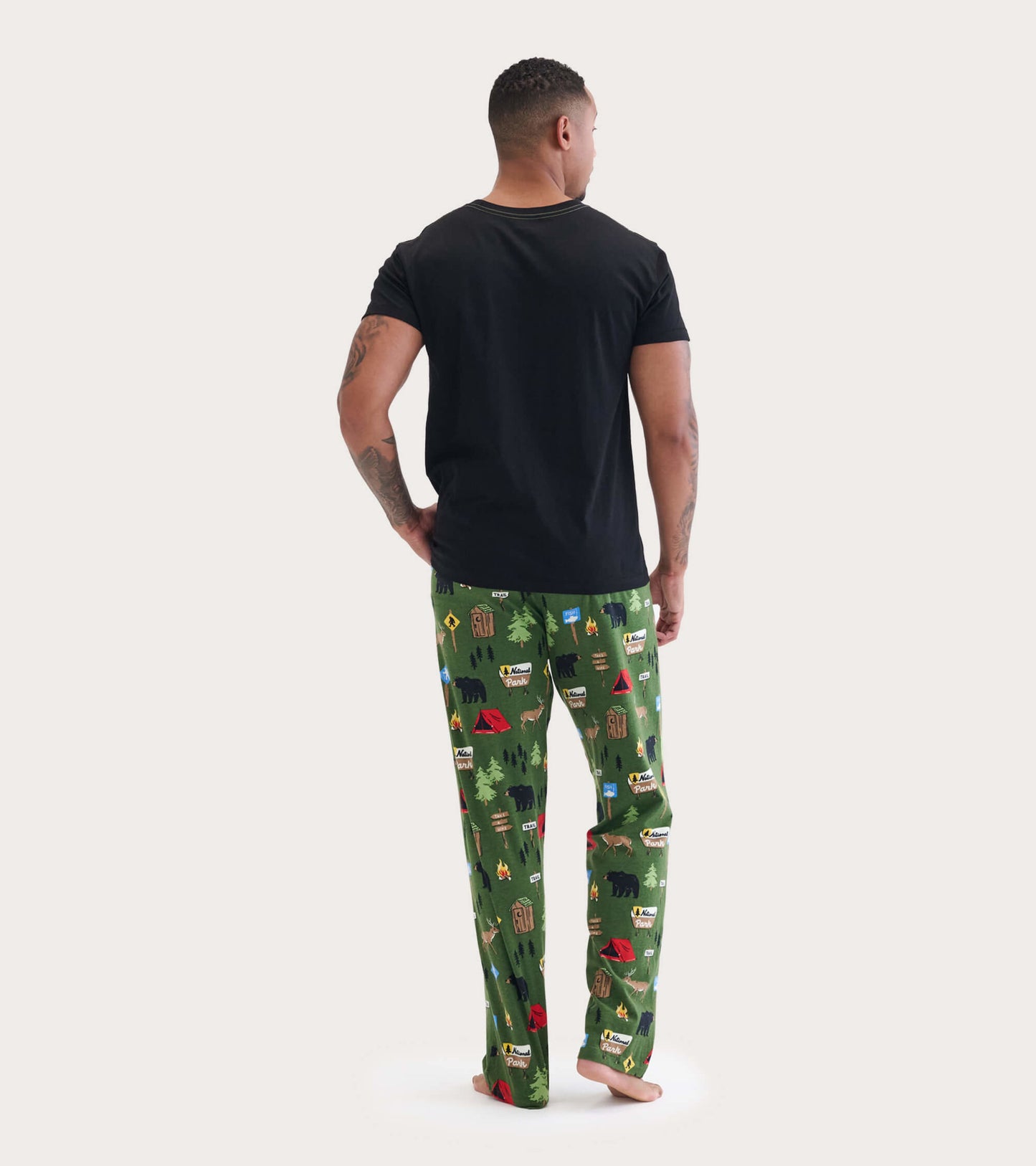 Black short-sleeve tee worn with patterned green pajama pants featuring camping and wildlife motifs, viewed from the back.