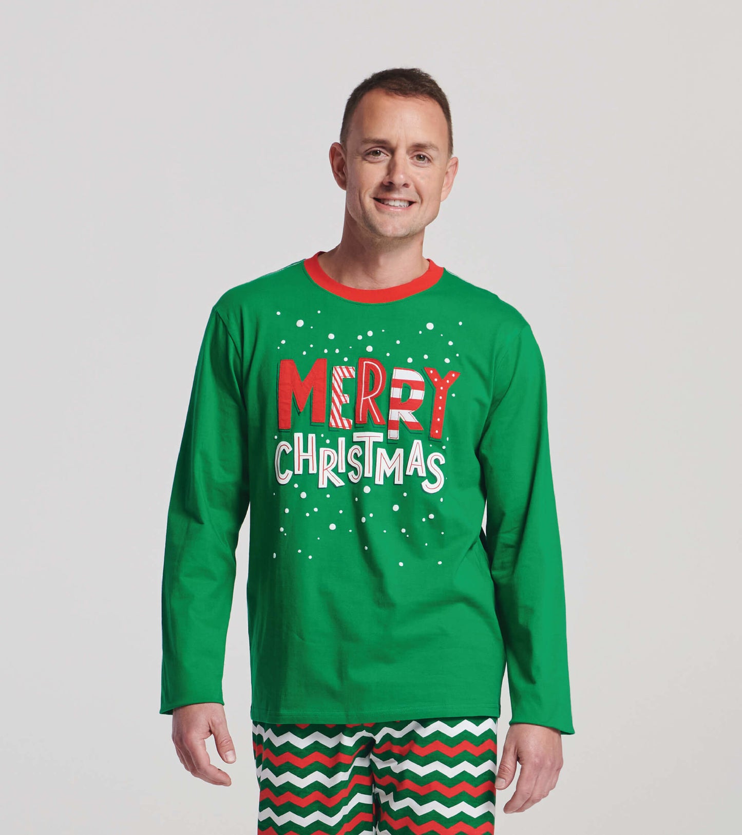 Green long sleeve t-shirt featuring "MERRY CHRISTMAS" graphic in red and white, worn with festive patterned pants.