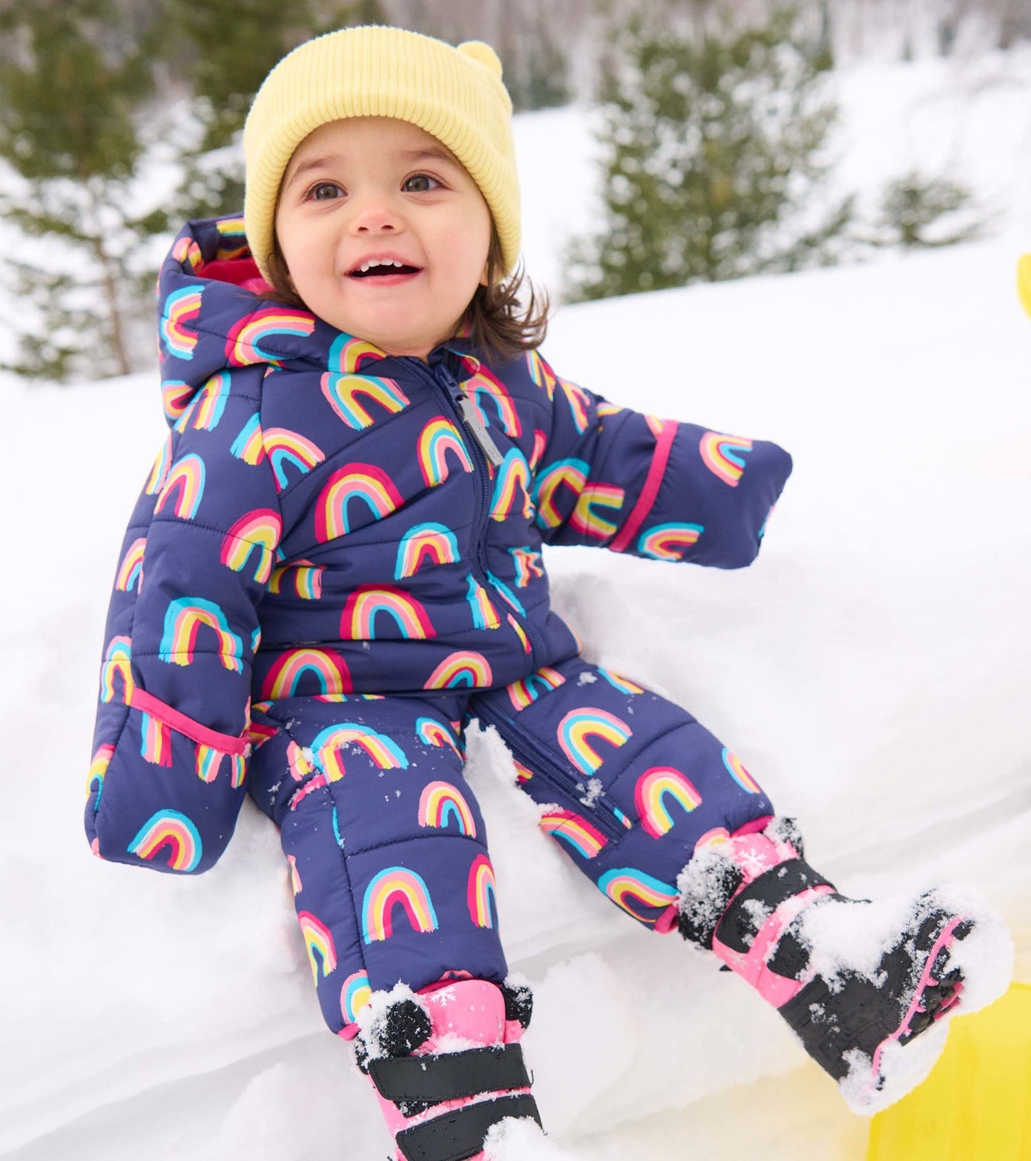 Colorful baby snowsuit featuring a rainbow pattern, paired with a yellow beanie, worn in a snowy outdoor setting.