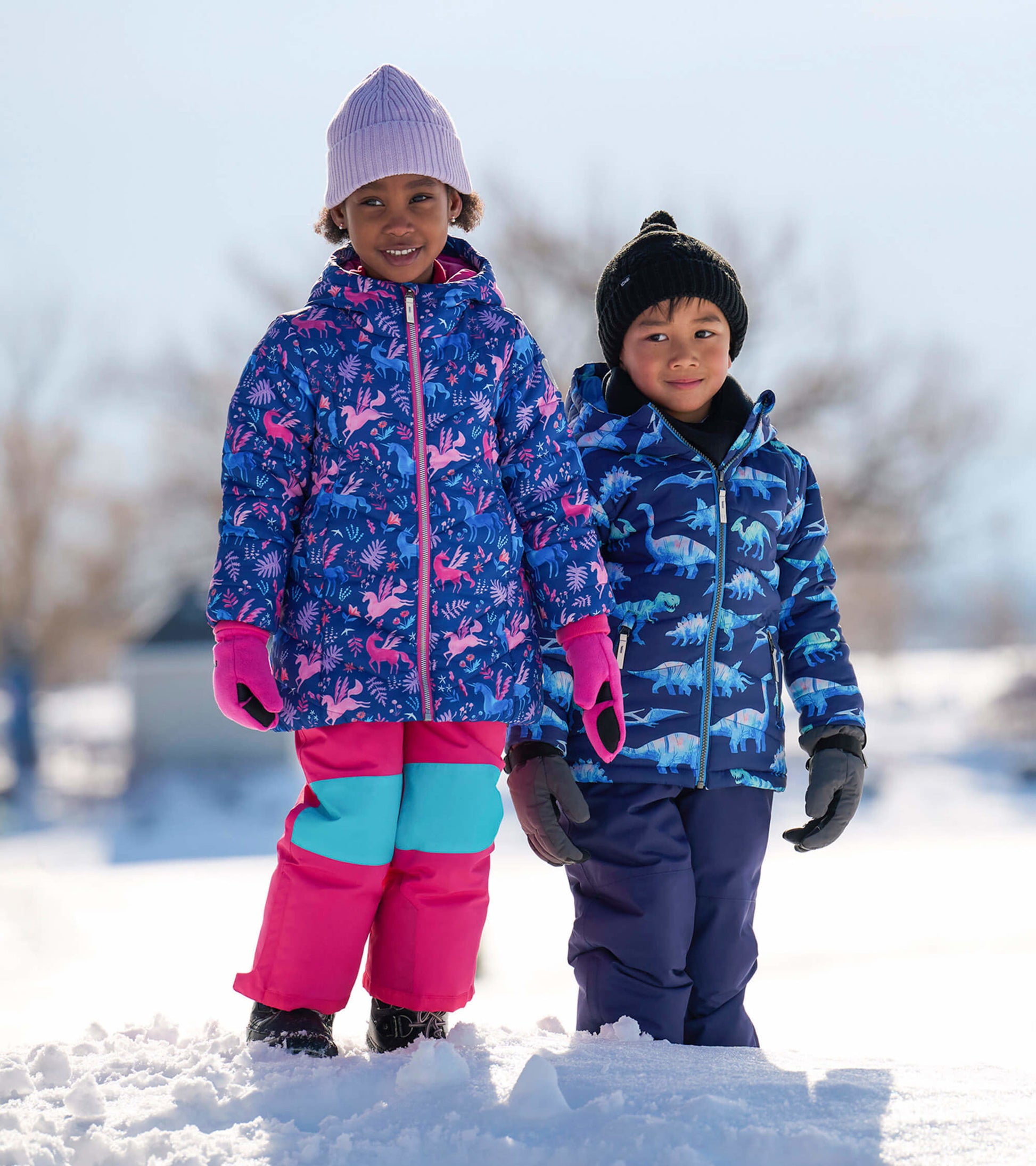 Colorful puffer jackets for kids, featuring a unicorn pattern and a dinosaur design, worn with snow pants in a snowy setting.