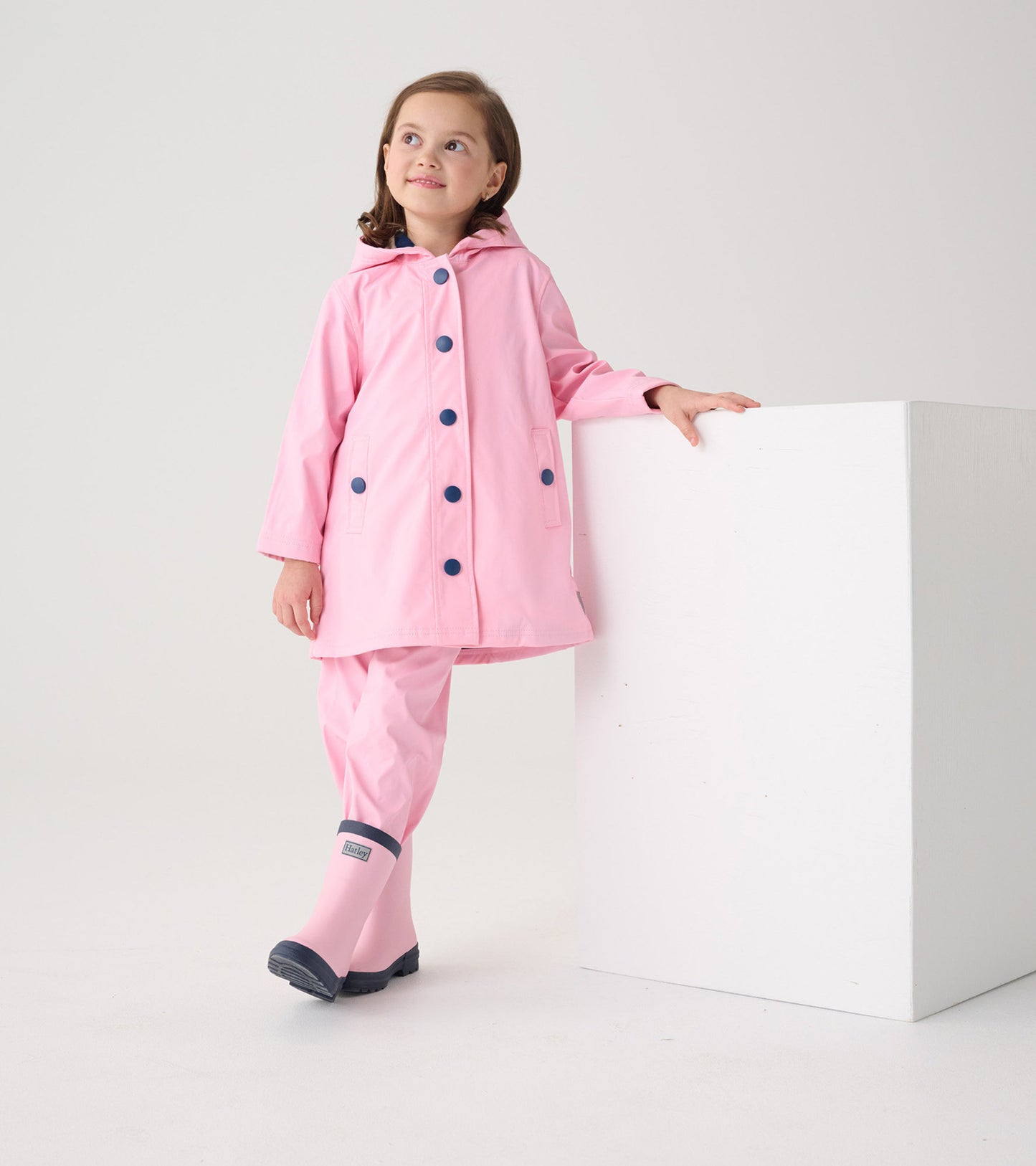 Pink button-up rain jacket with navy buttons and hood, worn by a girl, paired with matching pink rain pants and boots.