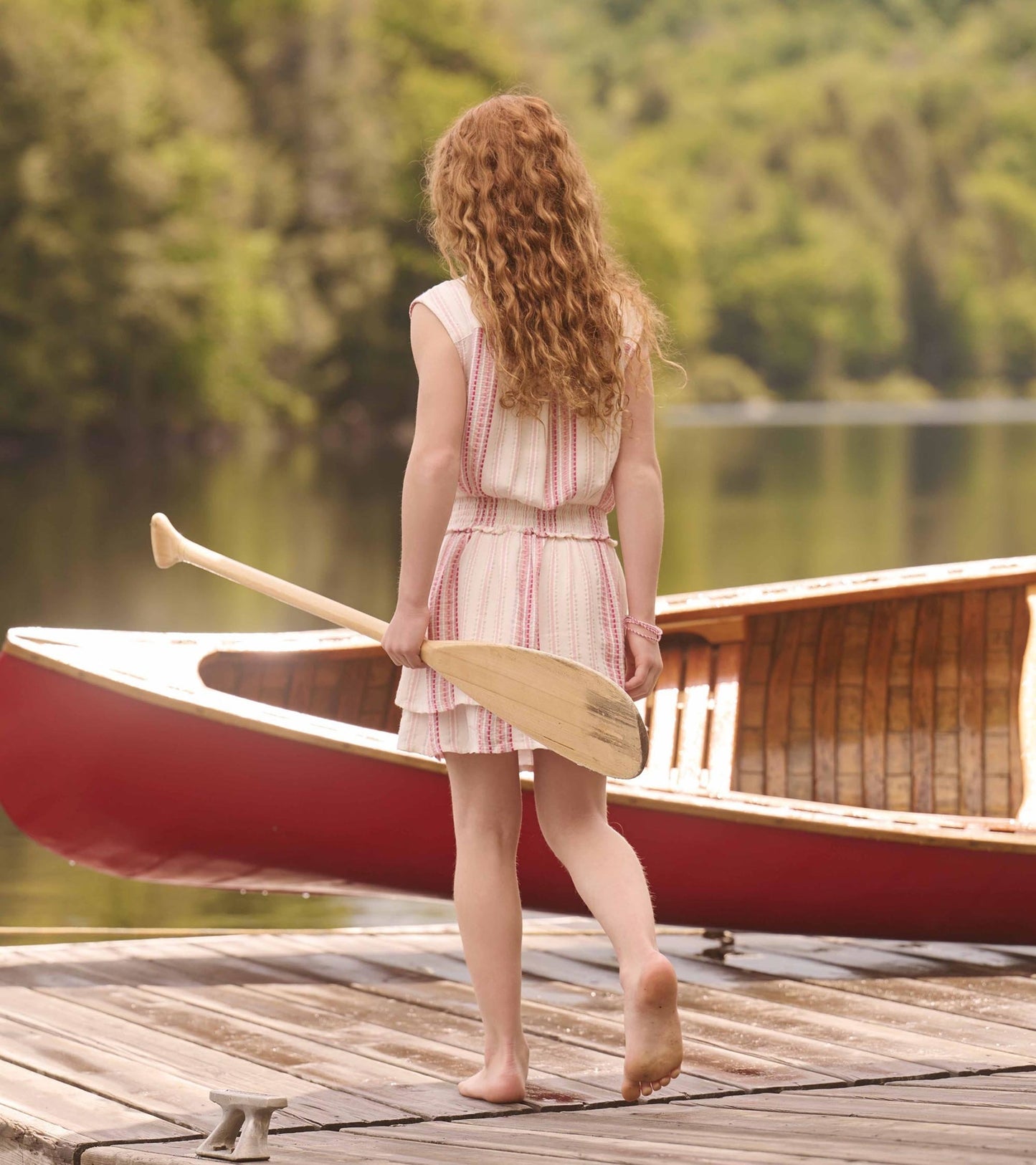 Girls' sleeveless dress with a smocked waist, featuring a pink and white stripe pattern, worn barefoot by a lake.