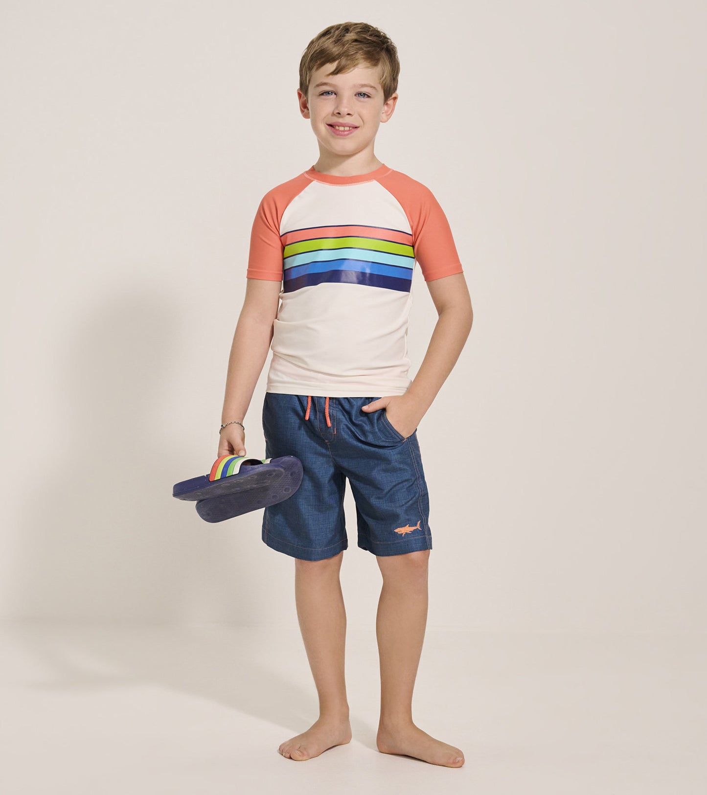 Short sleeve rashguard featuring orange sleeves and colorful horizontal stripes, paired with blue shorts and flip-flops.