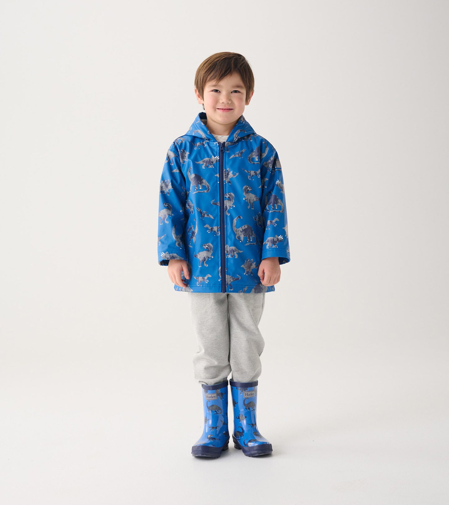 Blue zip-up rain jacket featuring a dinosaur pattern, worn by a child with gray pants and matching rain boots.