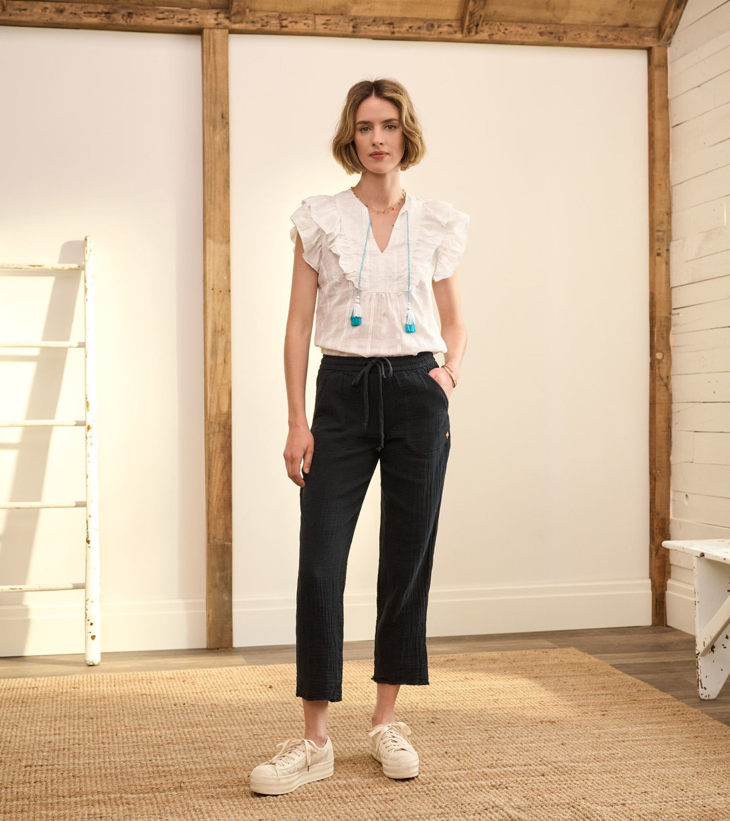 White ruffled blouse with a V-neck and blue tassels paired with dark plaid pants, worn with white sneakers.
