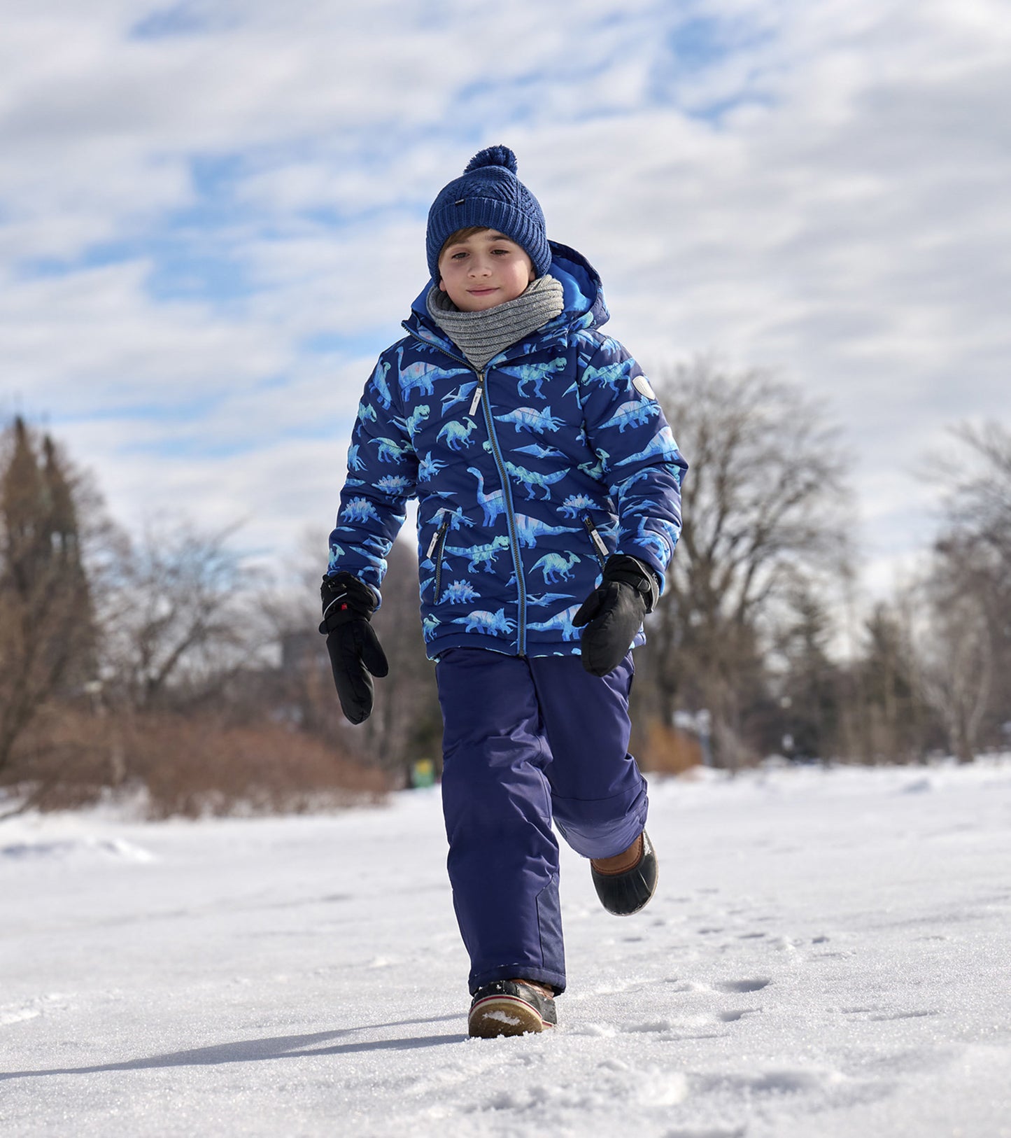 Blue puffer jacket with dinosaur prints, worn by a child in snow, paired with gloves and a knitted hat.