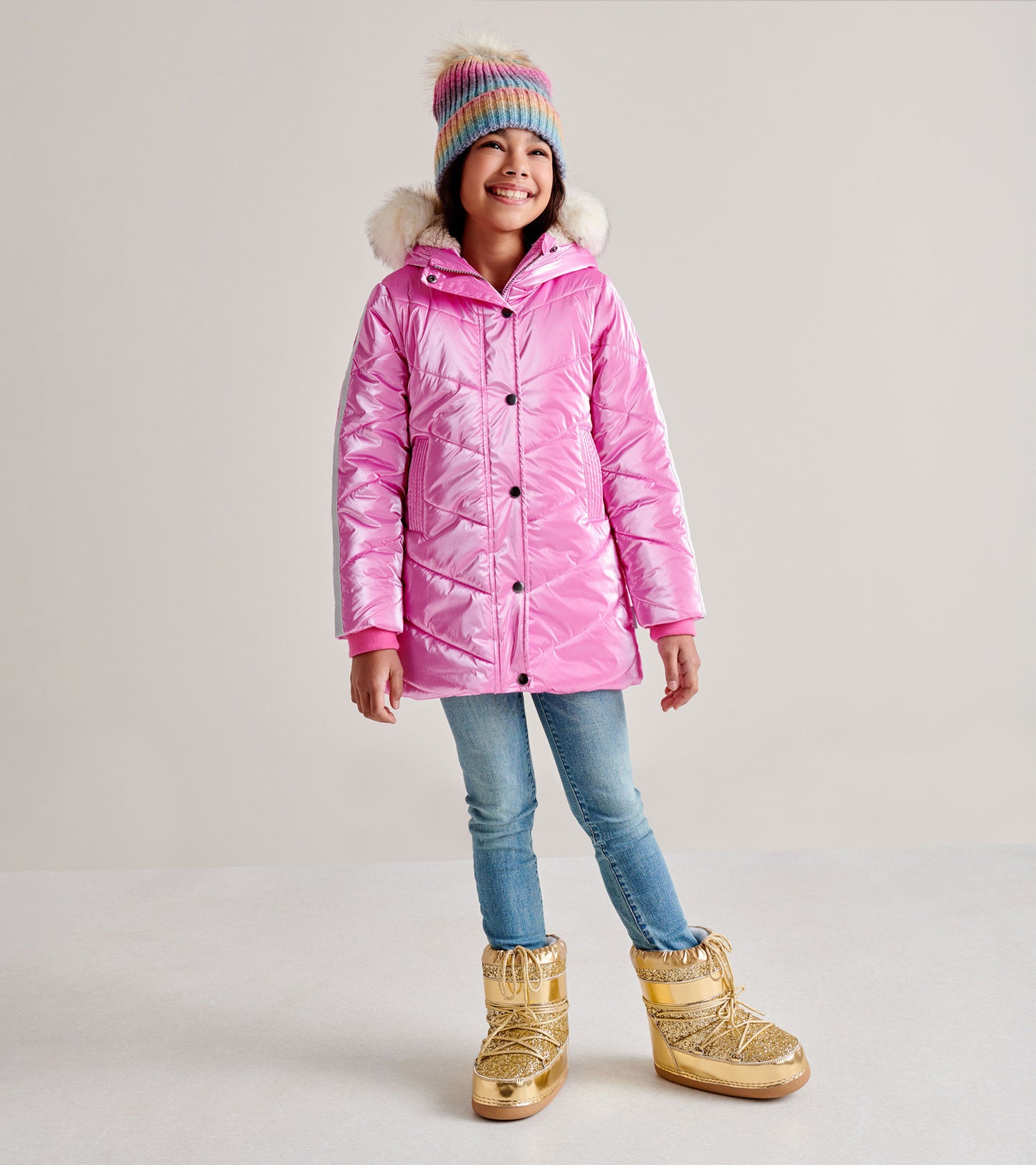 Shiny pink puffer jacket with a hood and faux fur trim, worn by a girl with gold winter boots and blue jeans.