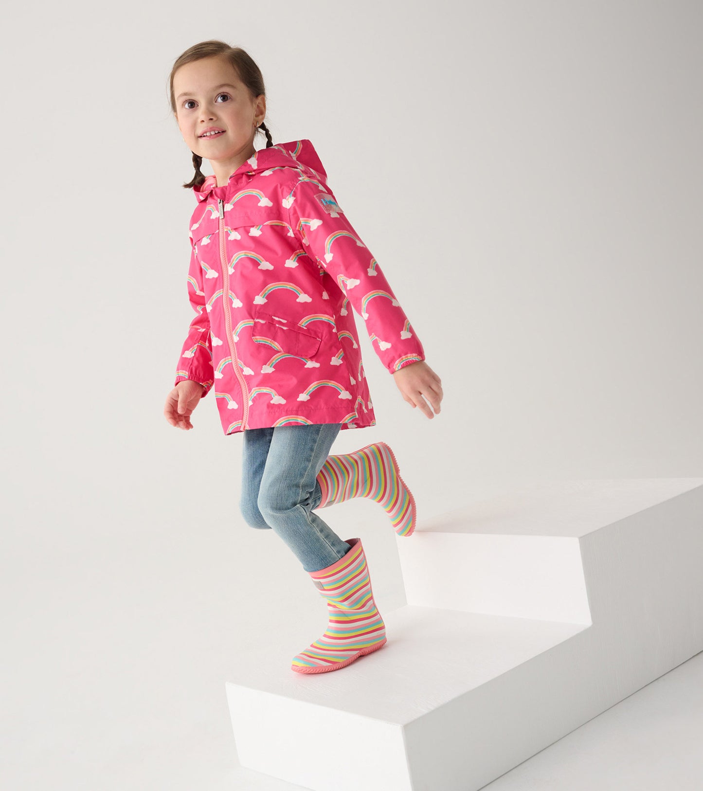 Lightweight pink rain jacket with rainbow pattern, hood, and zip-up front, worn by a girl on stairs with striped rain boots.
