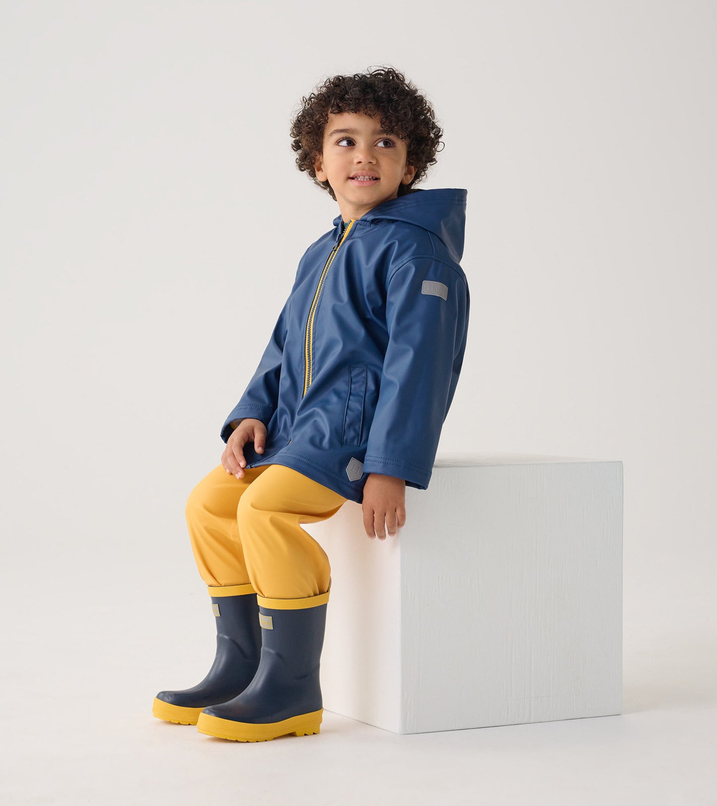 Navy zip-up rain jacket on a child, paired with yellow pants and navy rain boots, sitting on a white block.