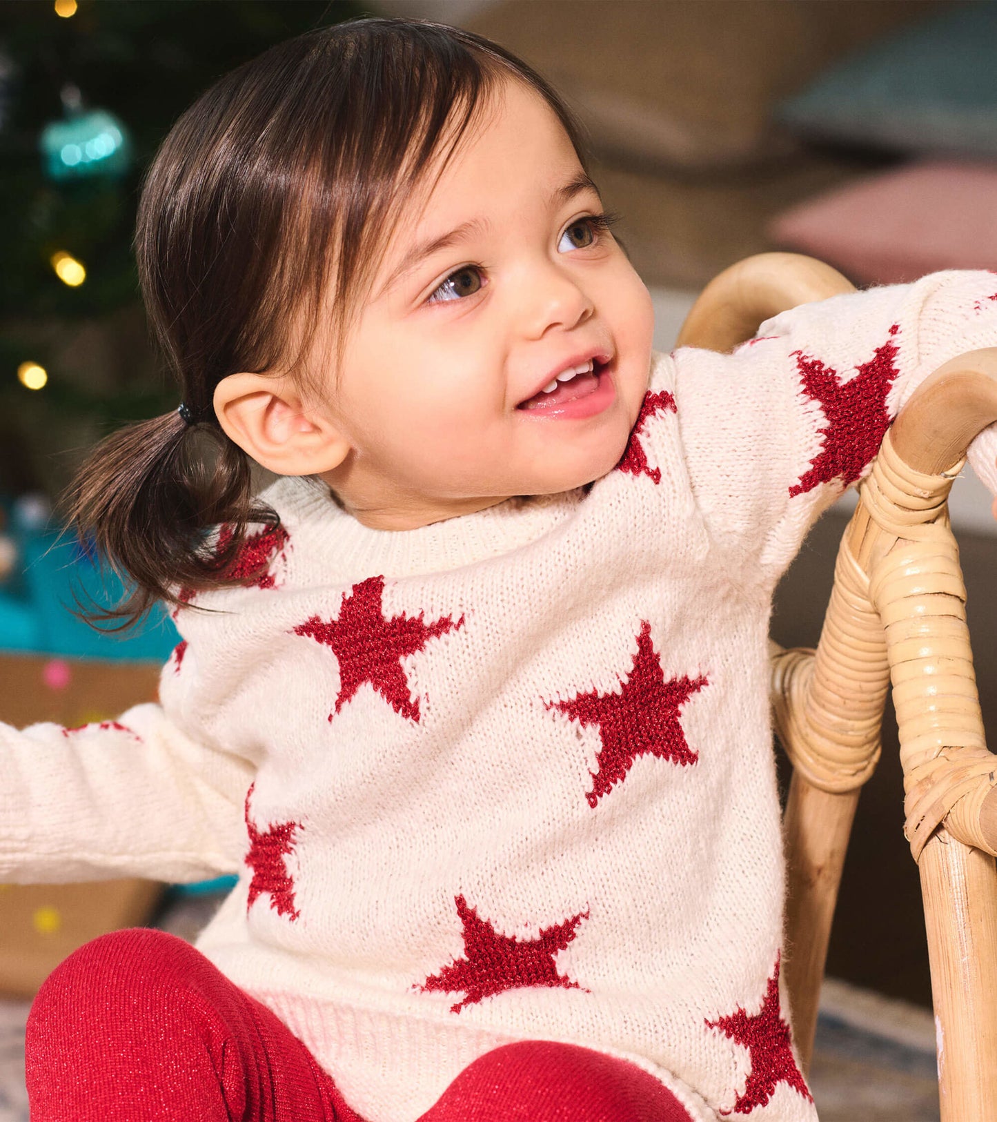 Cream sweater dress with red star pattern, worn by a child, paired with red leggings, sitting on a wicker chair.