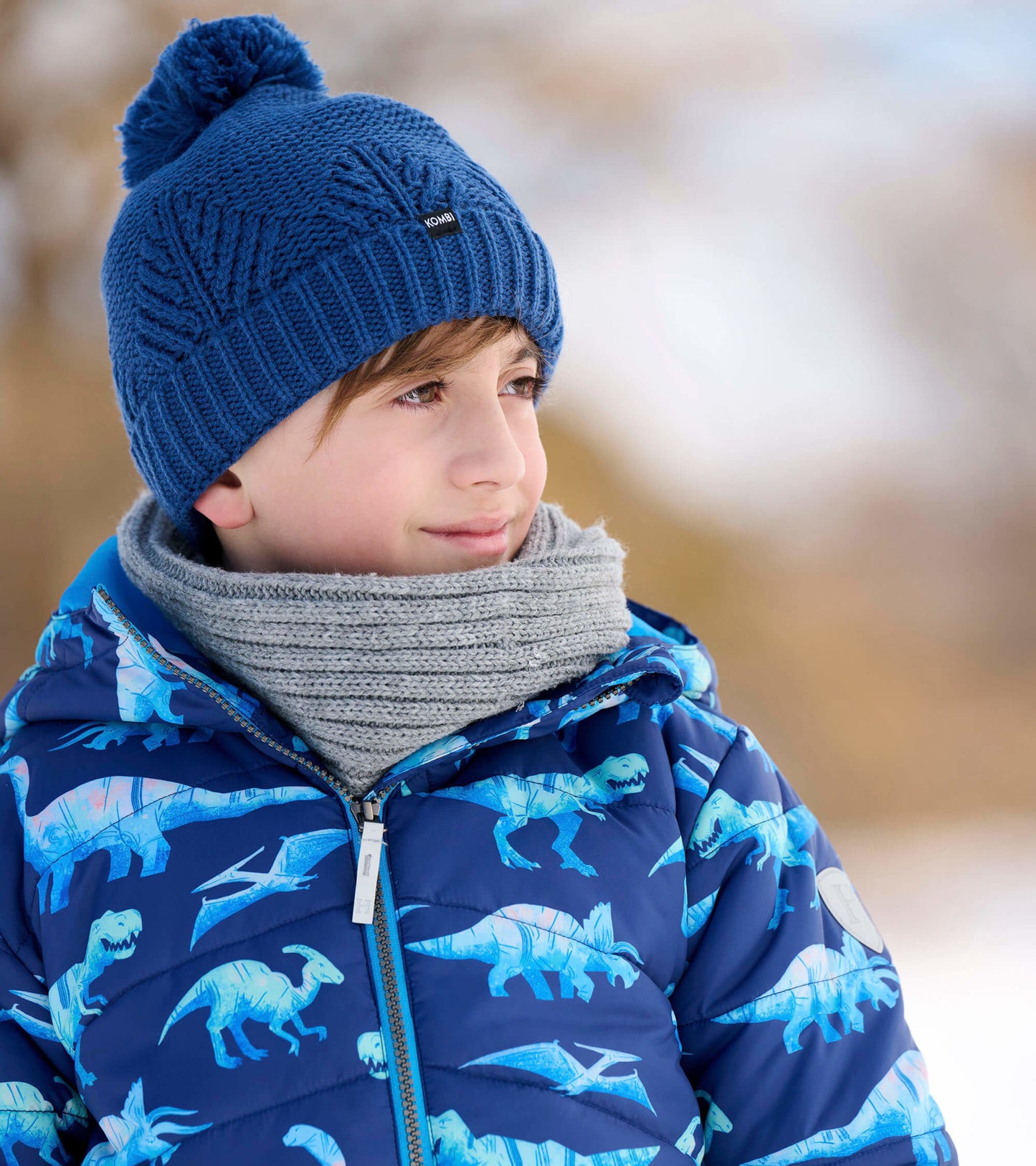 Blue puffer jacket featuring dinosaur patterns, worn by a child with a gray scarf and blue knit hat.