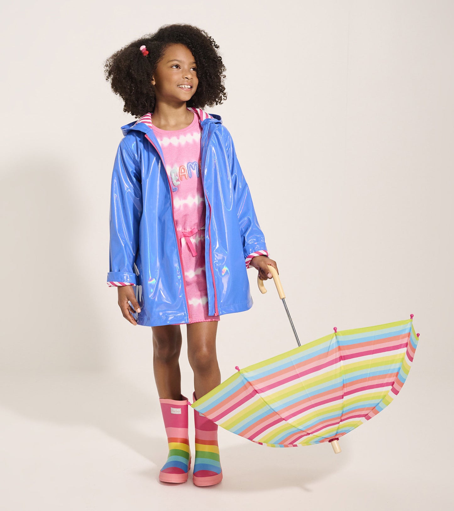 Blue glitter swing jacket with a hood, worn over a pink dress, paired with rainbow-striped umbrella and boots.