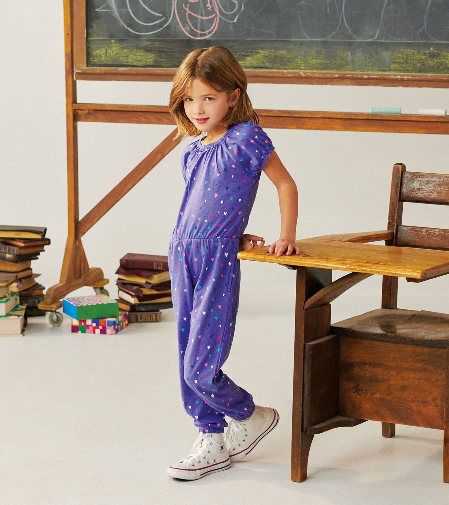 Purple jumpsuit with colorful heart pattern, short puff sleeves, and elastic waist, worn by a girl standing by a desk.