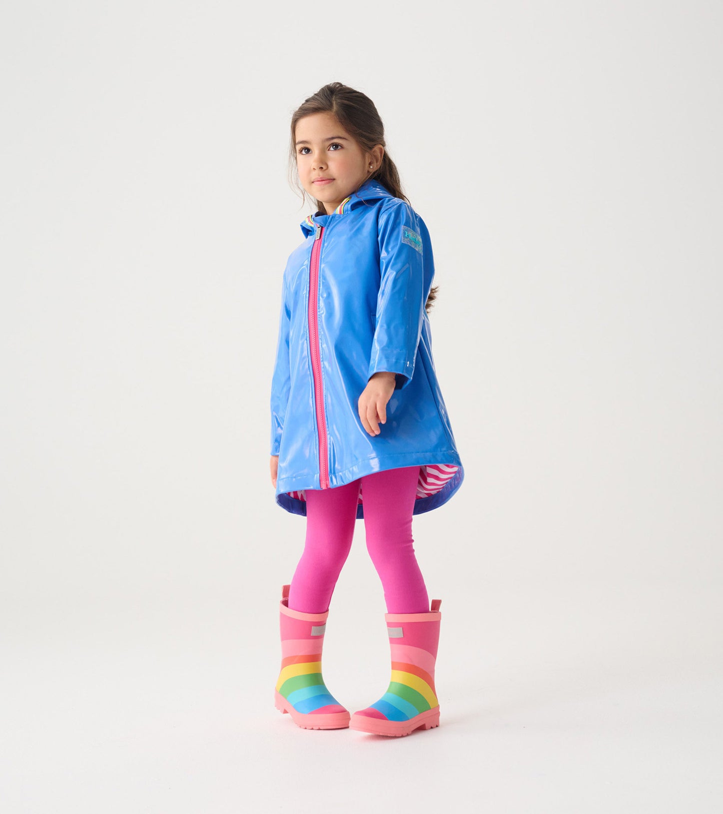 Blue glitter swing jacket with a hood, featuring a front zipper and a longer back hem, worn over pink leggings and rain boot…
