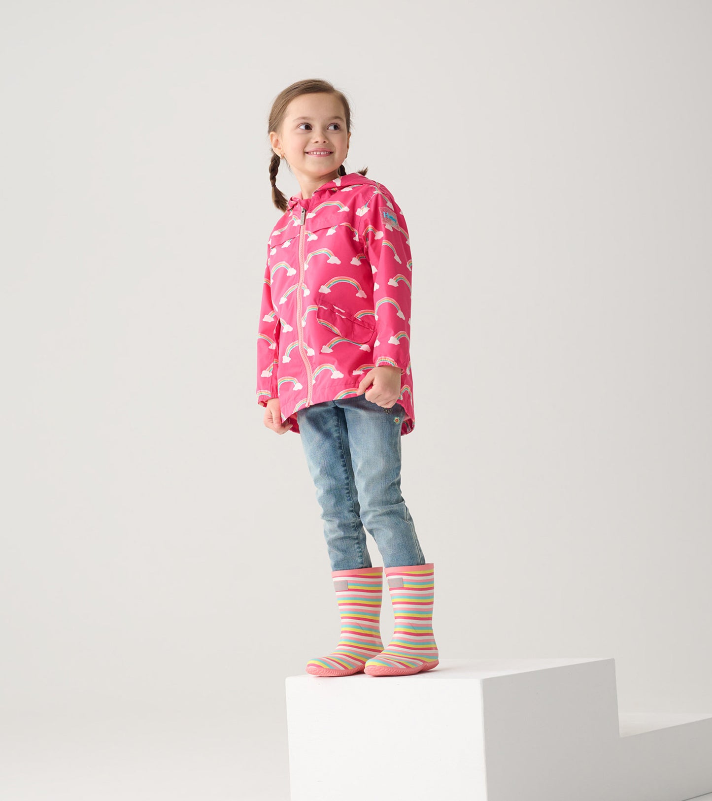 Lightweight pink rain jacket with rainbow pattern, paired with blue jeans and colorful striped rain boots.