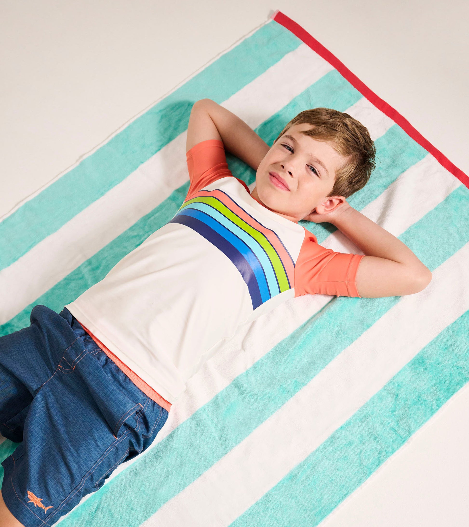 Short sleeve rashguard featuring orange sleeves and a colorful striped pattern, worn by a boy lying on a striped towel.