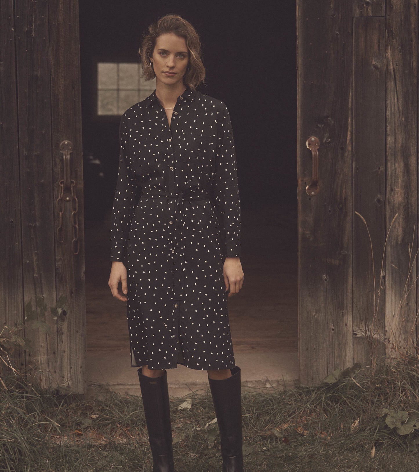 Black midi shirt dress with white polka dots, long sleeves, button front, and belted waist, styled with knee-high boots.