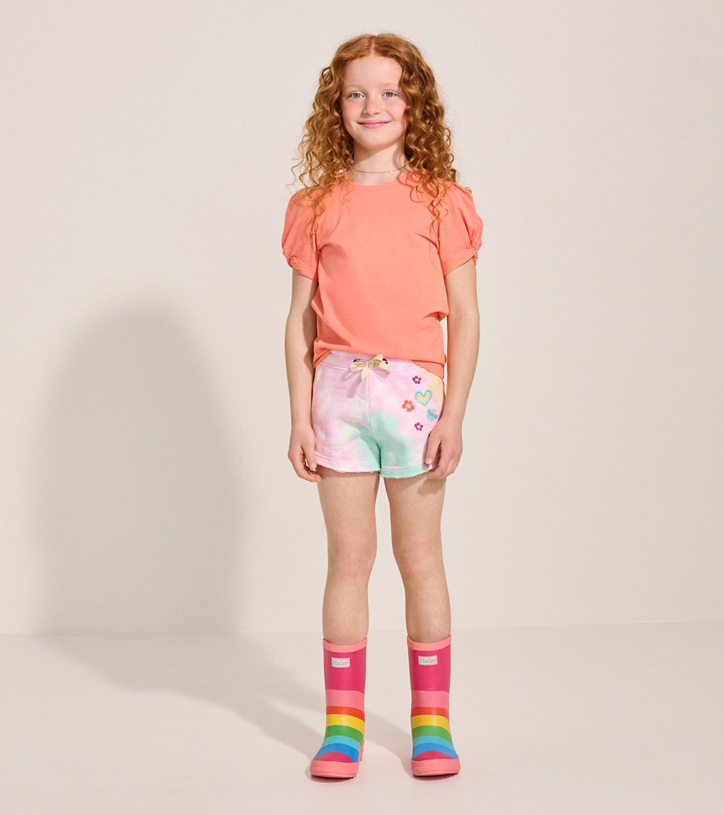 Colorful tie-dye pull-on shorts with floral designs, paired with a coral top and rainbow rain boots.